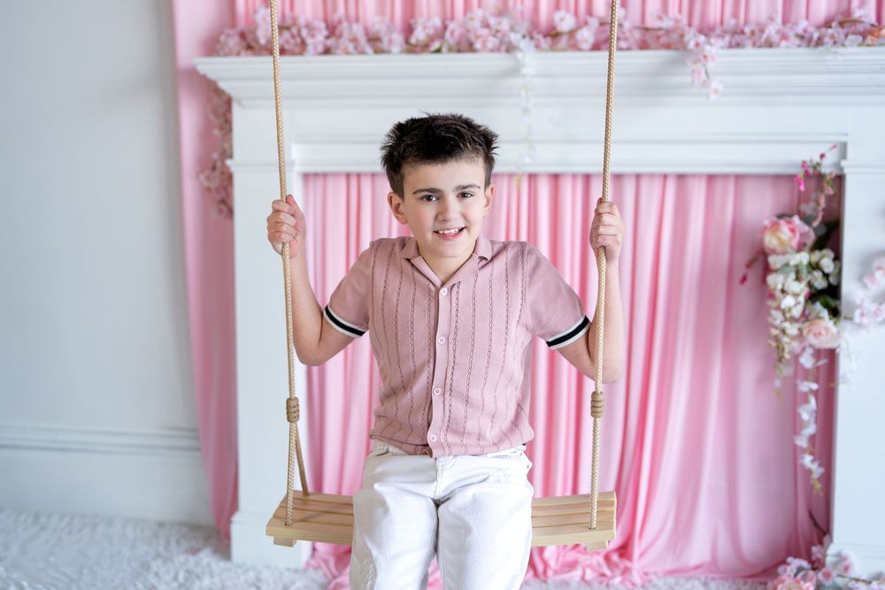 Happy young boy in pink shirt and white pants sitting on wooden swing smiling at camera in pink studio.
