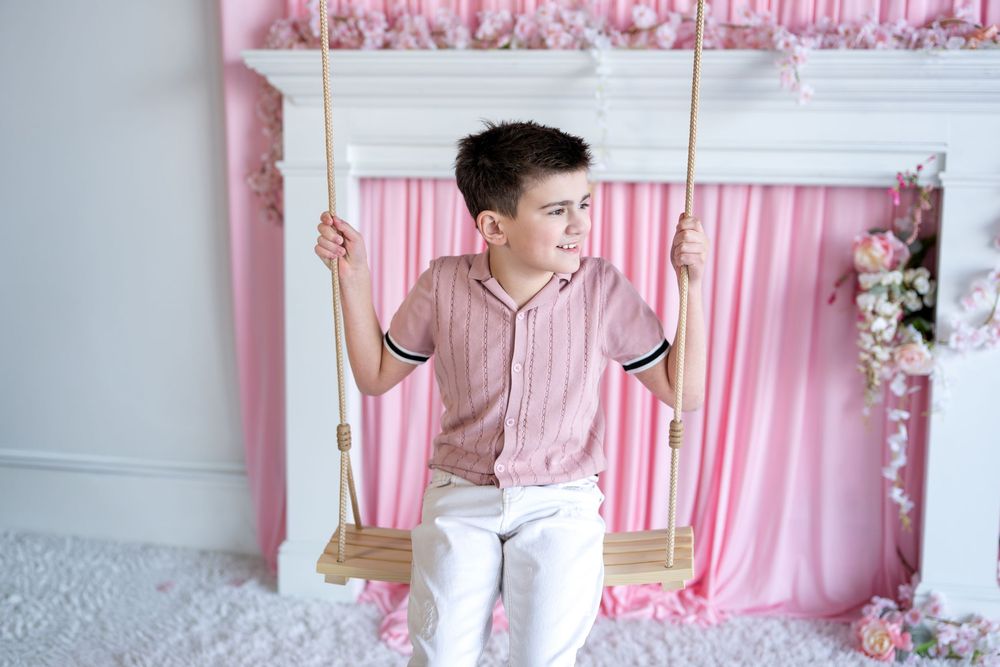 A boy in a pink striped shirt and white pants sits on a wooden swing against a pink curtain backdrop.