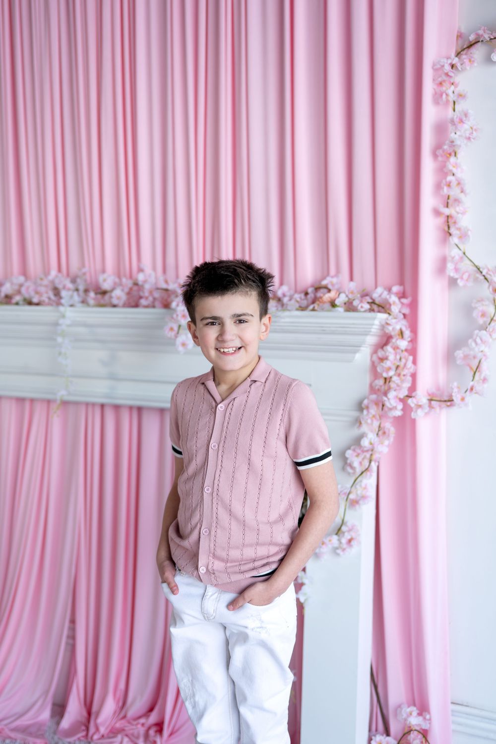 A smiling boy in a pink polo shirt and white pants stands with hands in pockets before a pink curtain backdrop.