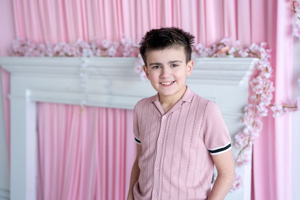 A cheerful boy in a pink polo shirt smiles in front of a white fireplace with pink curtains and flowers.