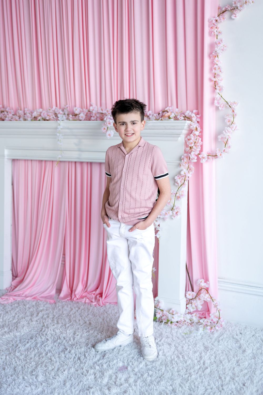A smiling boy in a pink shirt and white pants poses in front of a pink curtain with floral decorations.