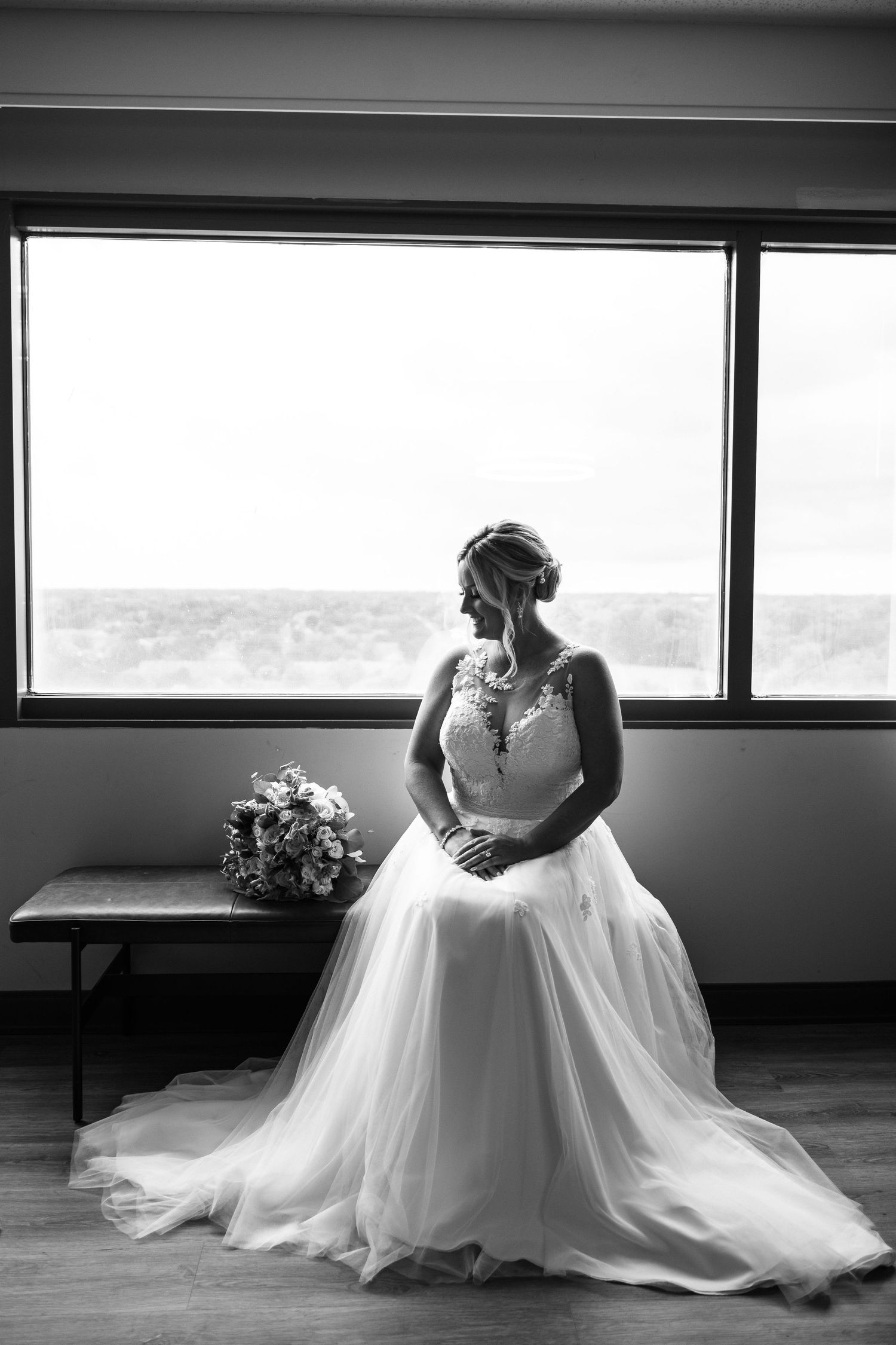 A bride in a flowing white wedding gown sits gracefully by a window in a series of elegant black and white portraits.