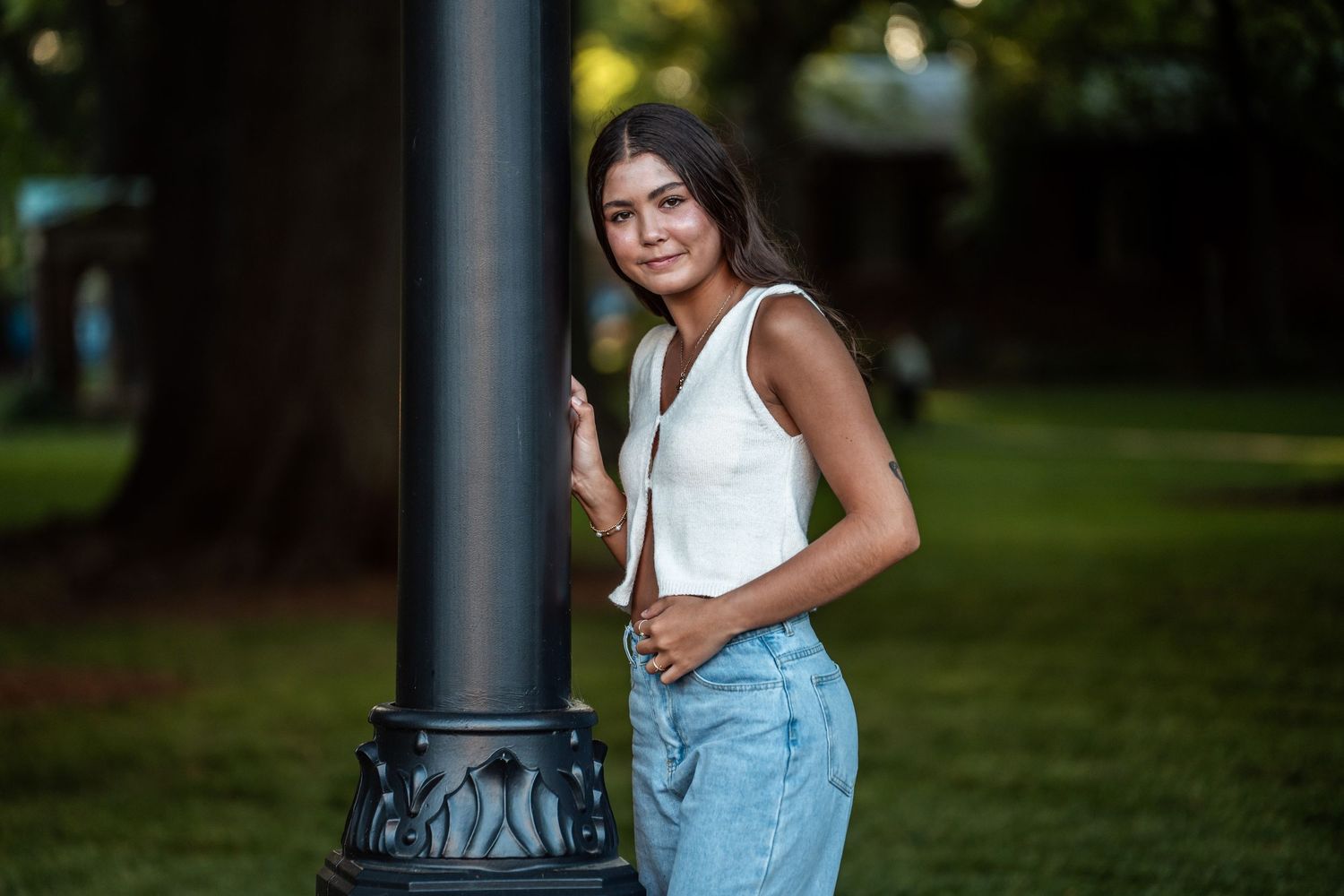Woman in white tank top and denim overalls poses against dark pole outdoors with blurred green background.