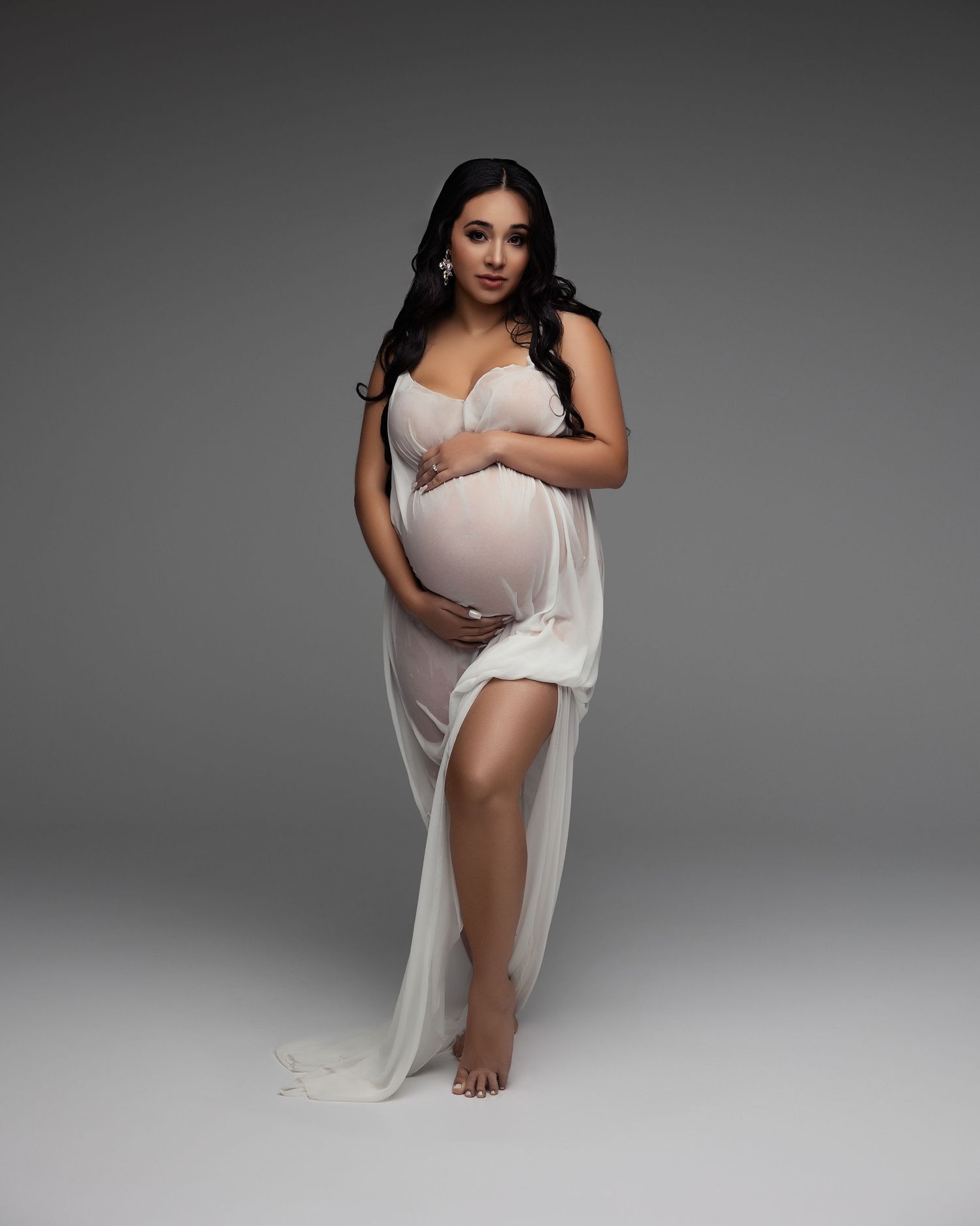 Elegant maternity portrait in white flowing dress against grey backdrop showcasing pregnancy silhouette.