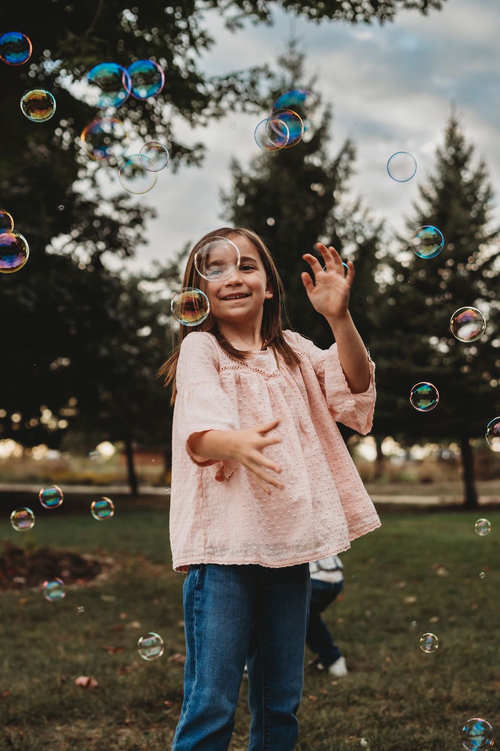 A child in a pink top plays with soap bubbles in a park setting. Family sessions