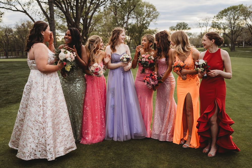 A prom party wearing vibrant dresses in different colors poses together on a lawn.