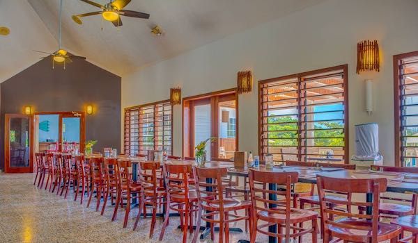 Spacious dining room with wooden chairs and large windows overlooking tropical landscape.