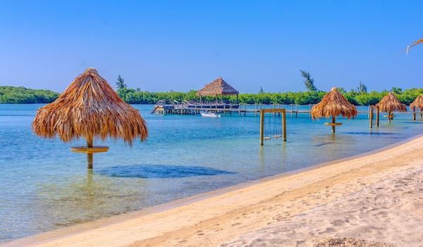 Palm-thatched umbrellas line a pristine beach with clear turquoise waters and overwater bungalows in the distance.
