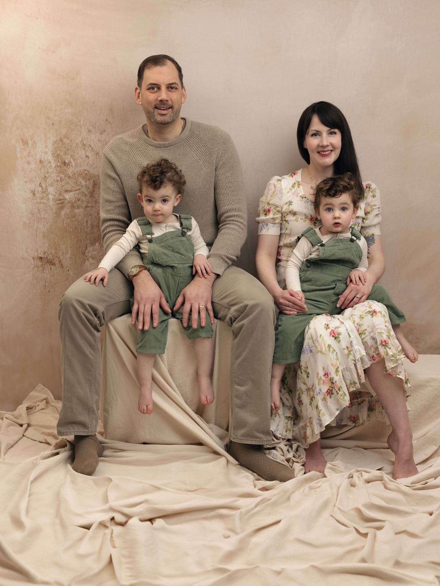 Family portrait with parents and twin toddlers in matching green overalls against beige backdrop and fabric.