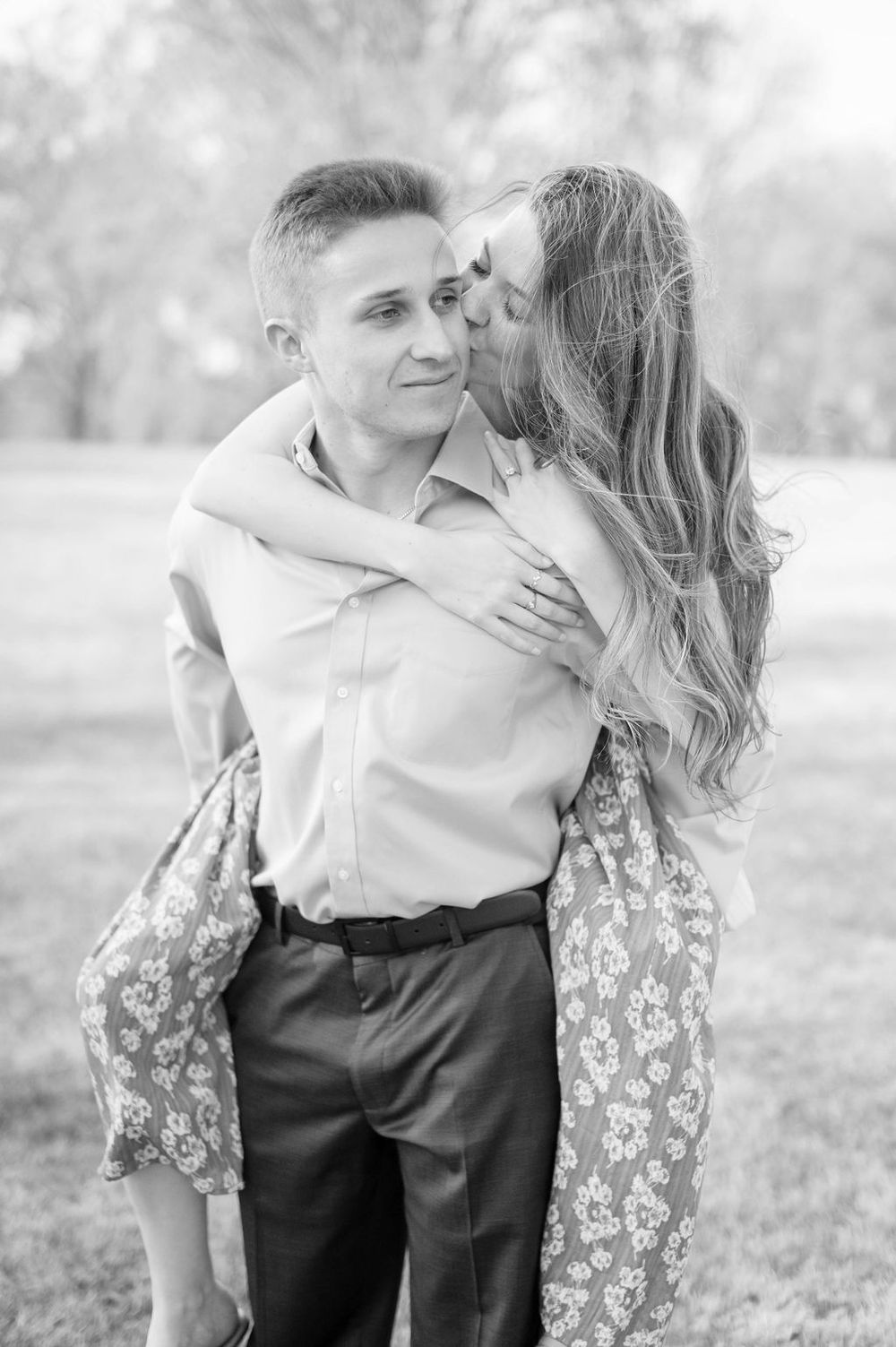 A romantic black and white portrait of a couple embracing outdoors, with her wearing a floral dress and him in a dress shirt.