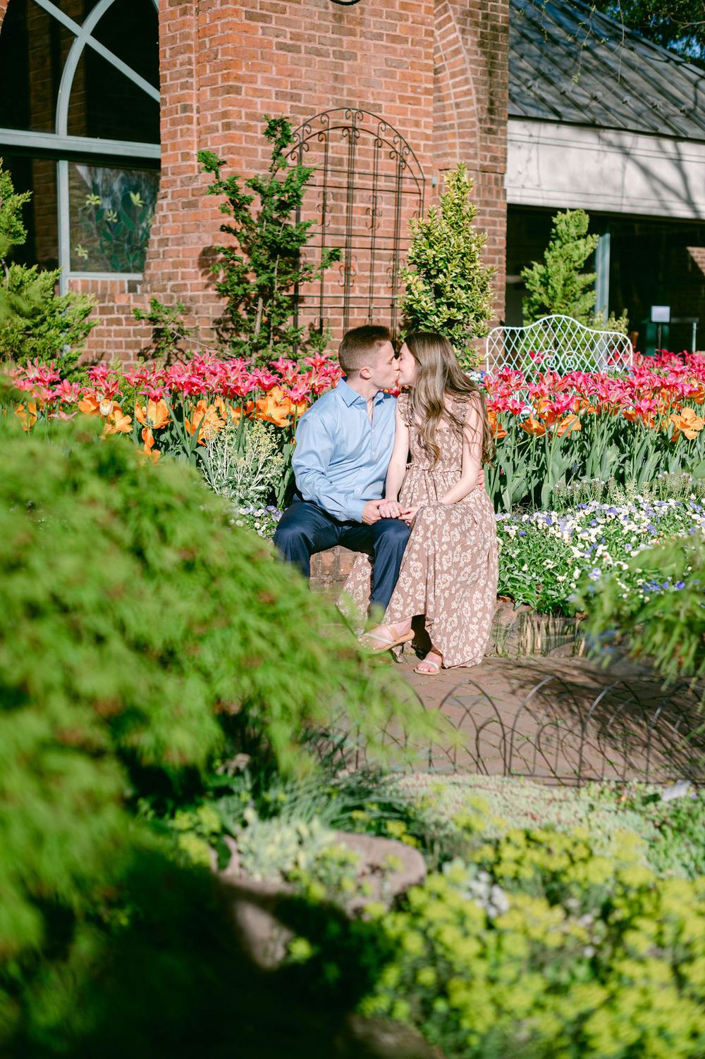 A couple shares a romantic moment in a garden with blooming tulips and brick building in the background.
