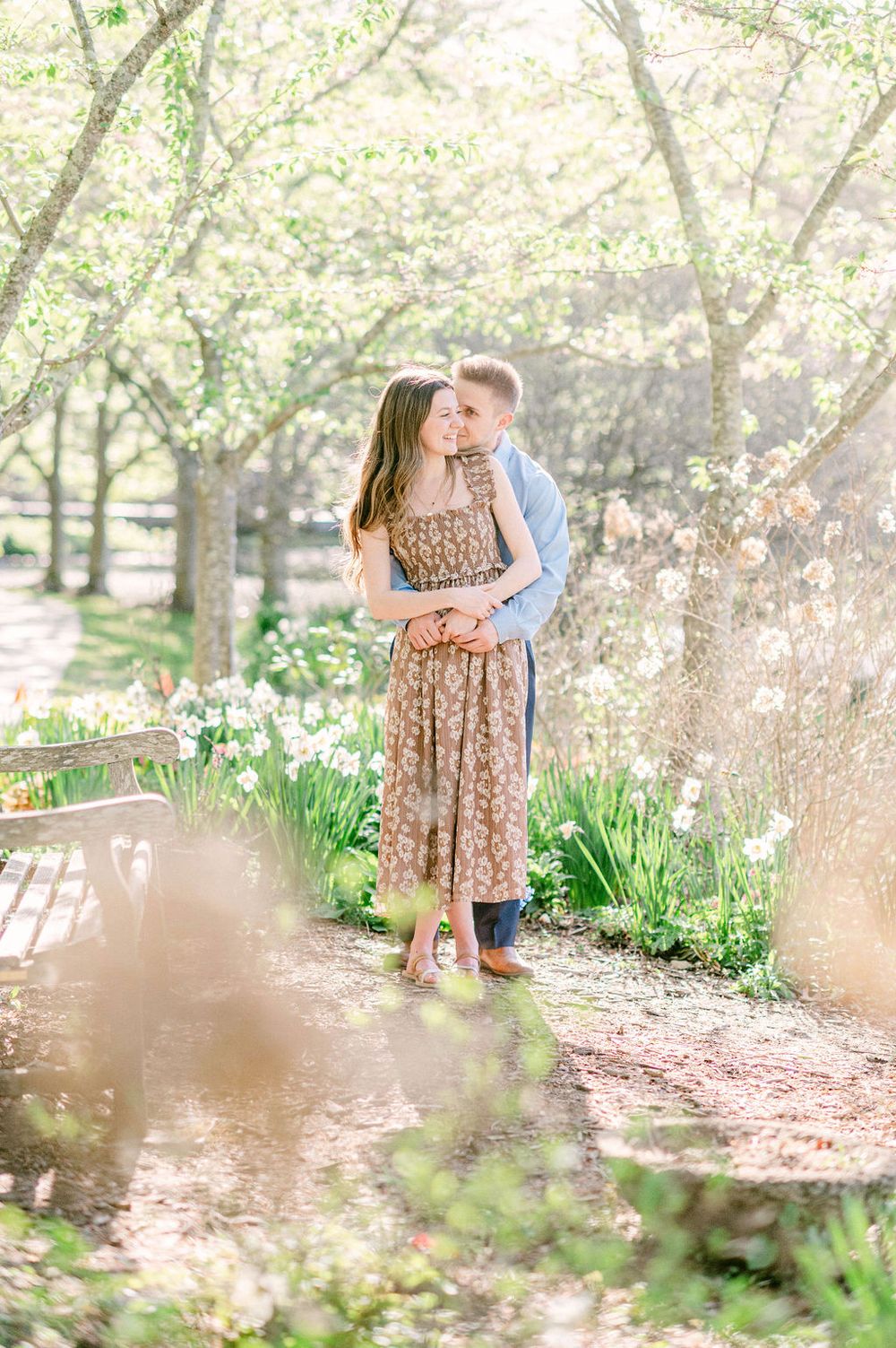 A couple embraces on a garden path surrounded by blooming spring trees and daffodils in soft natural light.