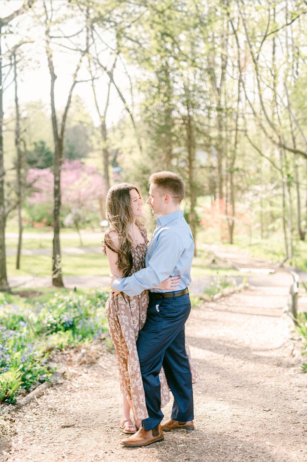 Two young children in matching outfits play together on a wooded path surrounded by spring flowers and trees.