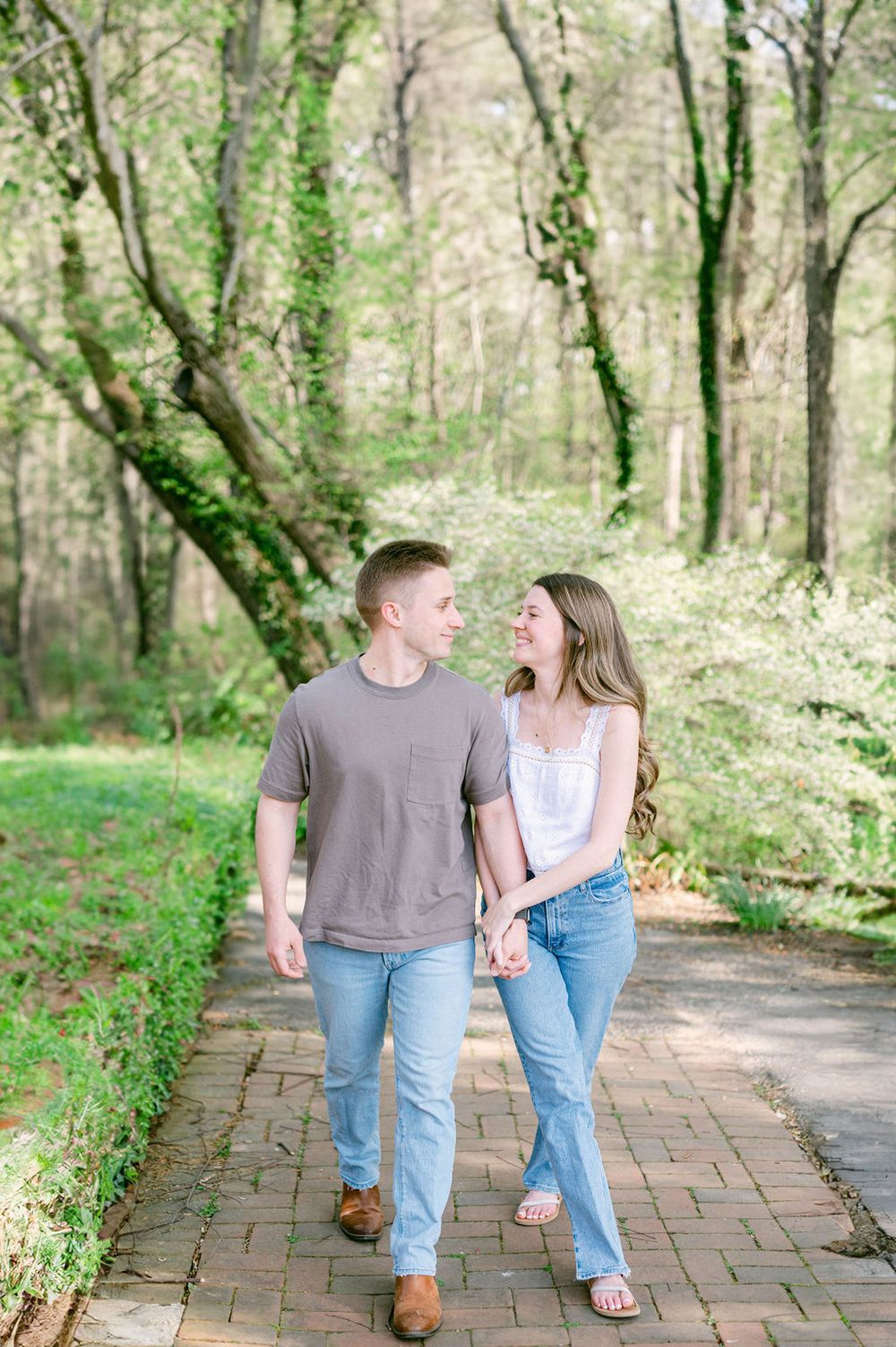 A couple walking hand in hand along a scenic brick path surrounded by lush green trees in a forest setting.
