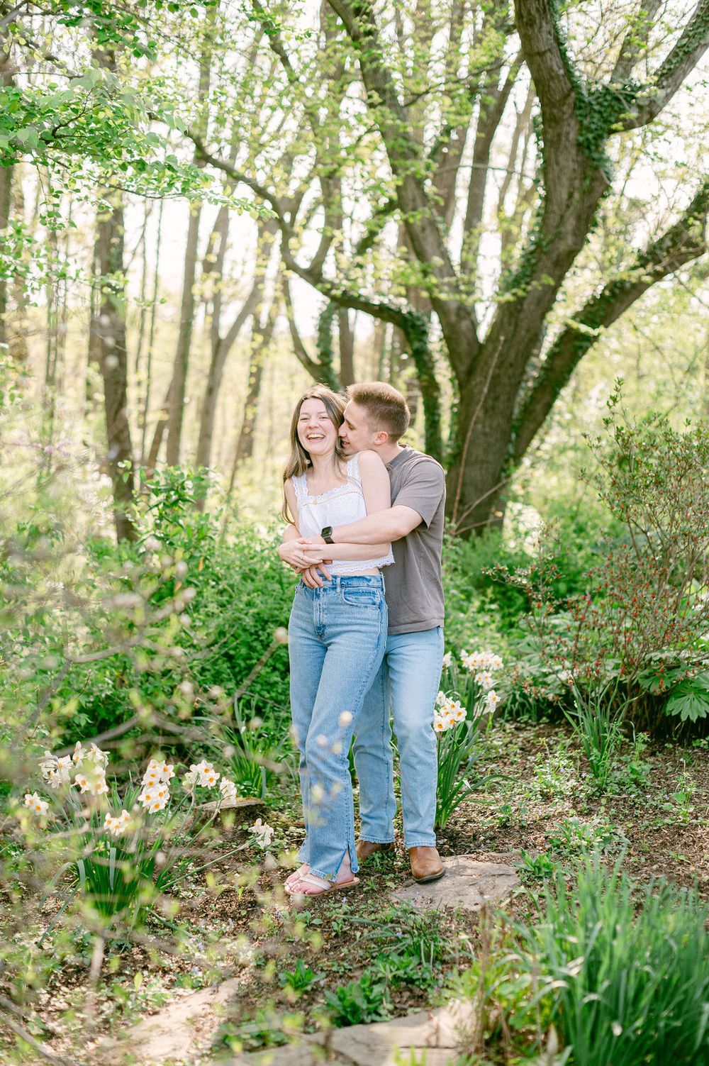 A couple shares intimate moments in a sunlit forest setting with lush greenery and spring flowers surrounding them.
