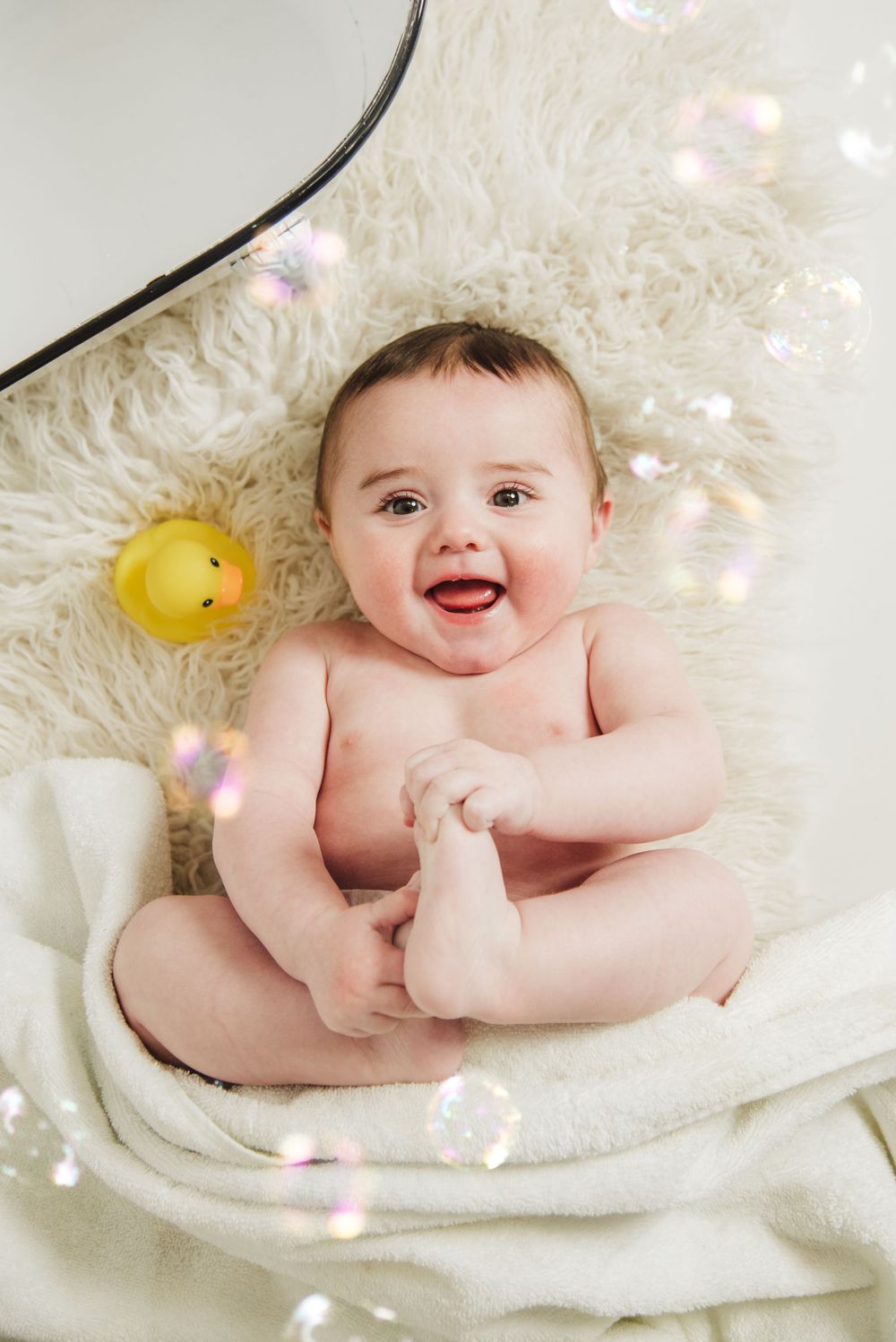 A joyful baby lies on a white fluffy blanket with sparkling bubbles and a yellow rubber duck toy in the scene.