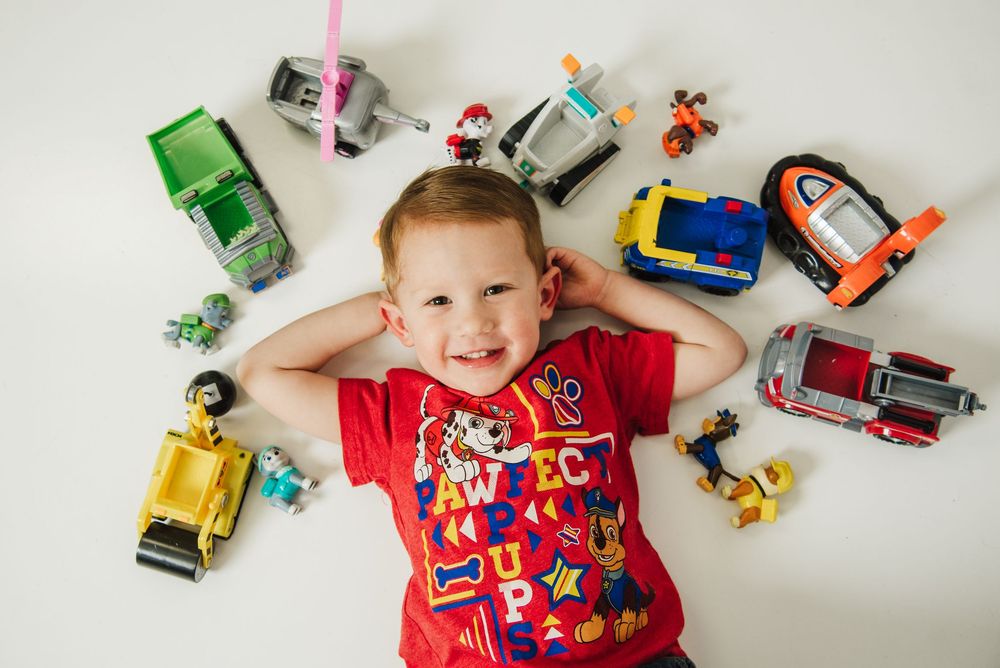 A child lies on a white surface surrounded by colorful toy cars and vehicles arranged in a circular pattern.