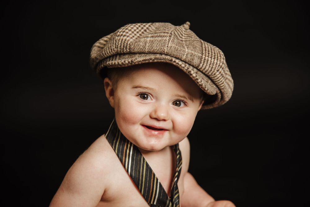 A baby wearing a brown tweed newsboy cap smiles sweetly at the camera.