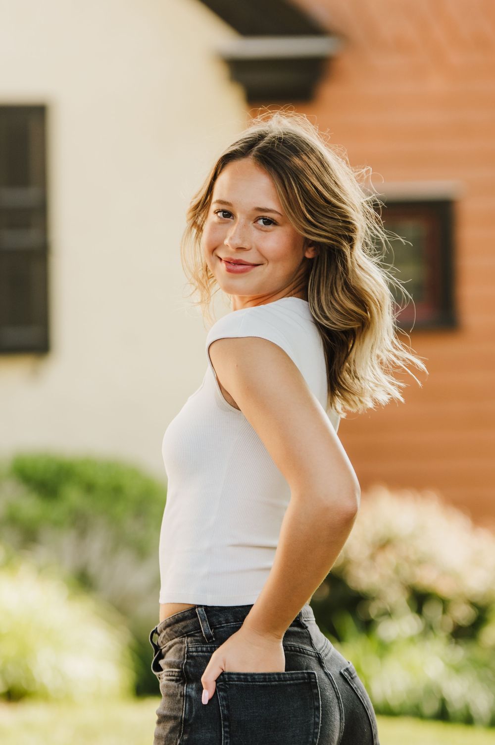 A person in a white tank top and jeans twirls and poses playfully outdoors against a brick wall background.