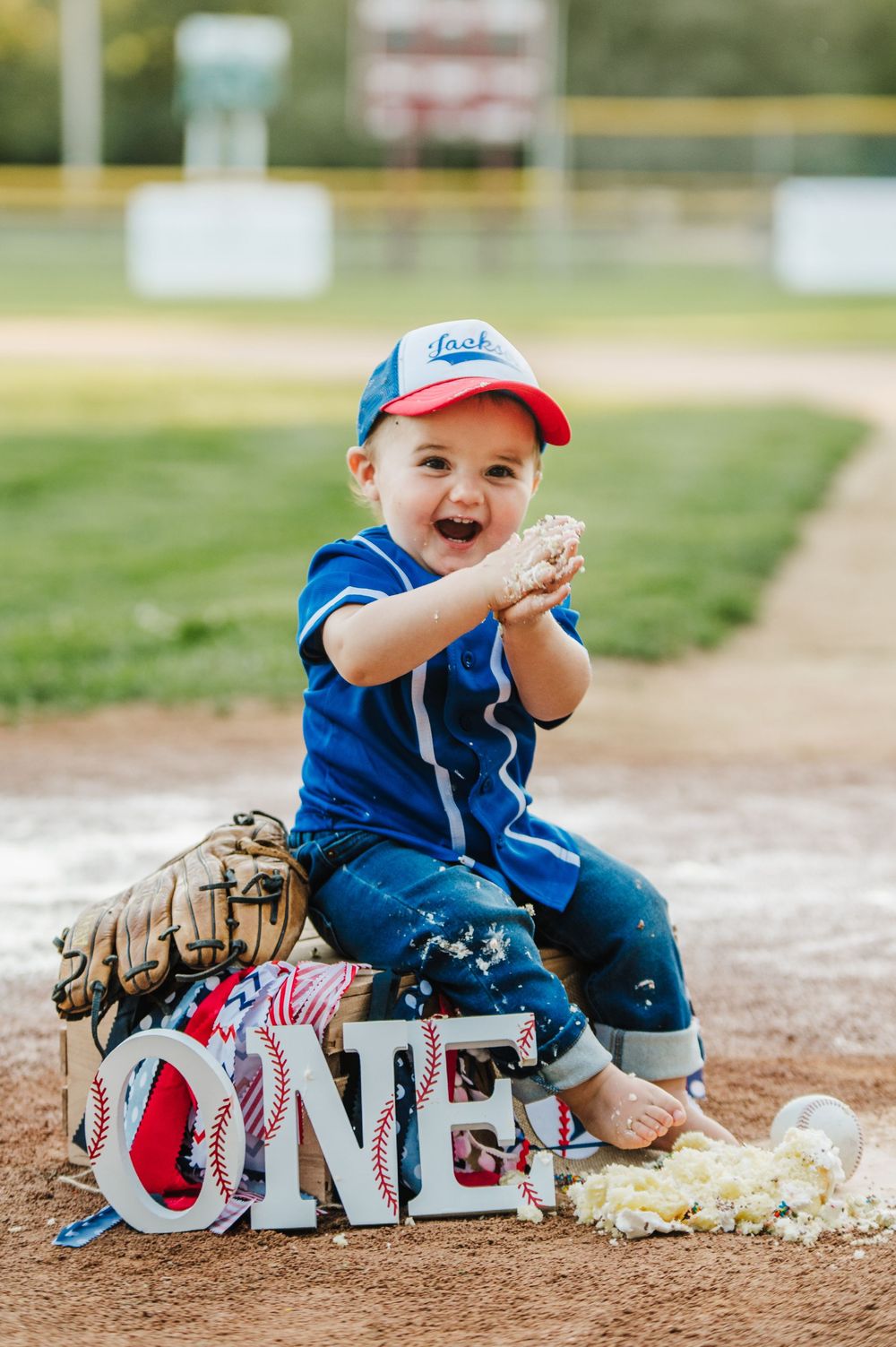 A small child in baseball attire sits on decorative letters spelling 'ONE' with a baseball glove on a baseball field.