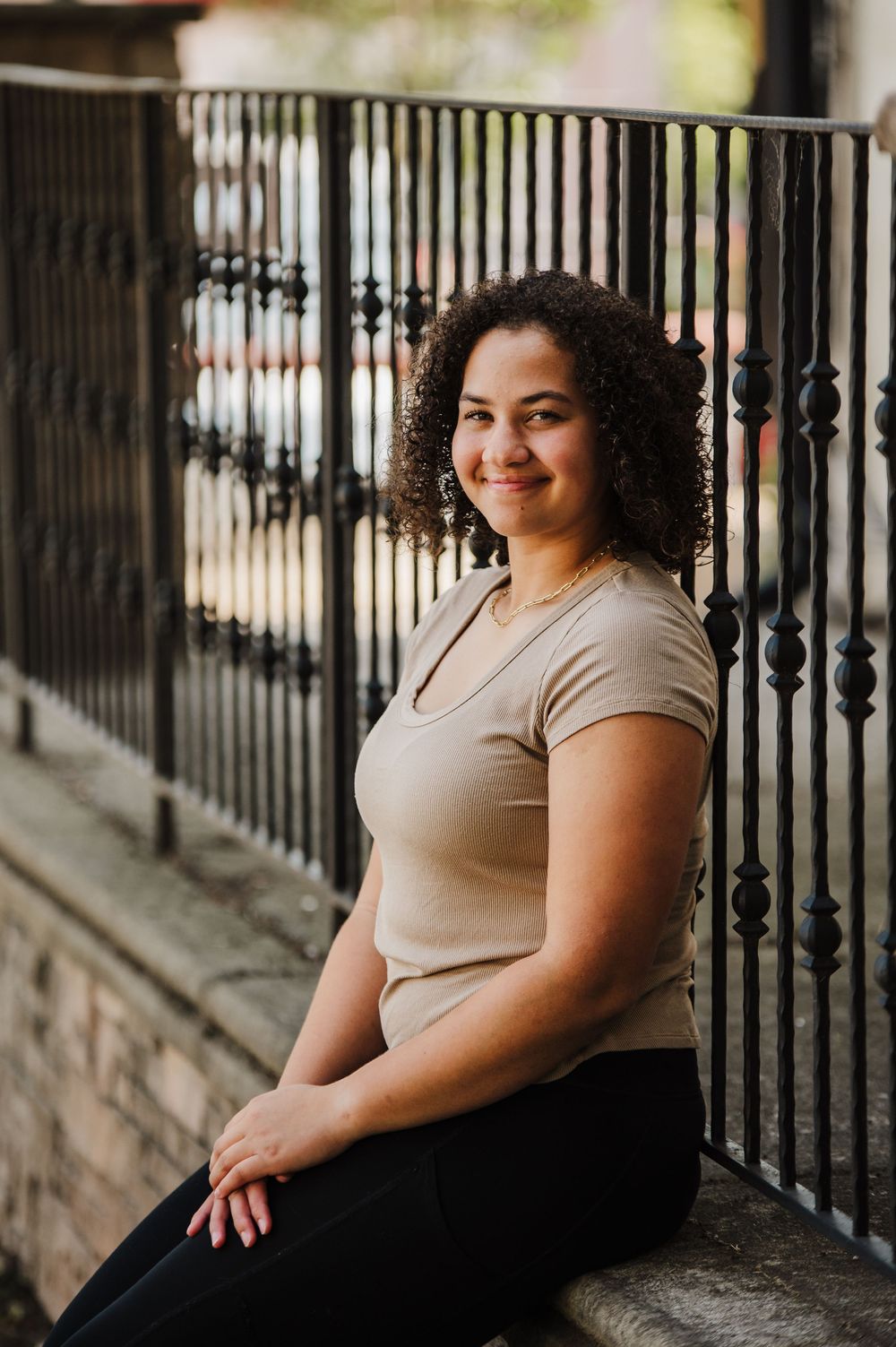 A curly-haired woman in a beige t-shirt and black pants poses outdoors against an iron railing.