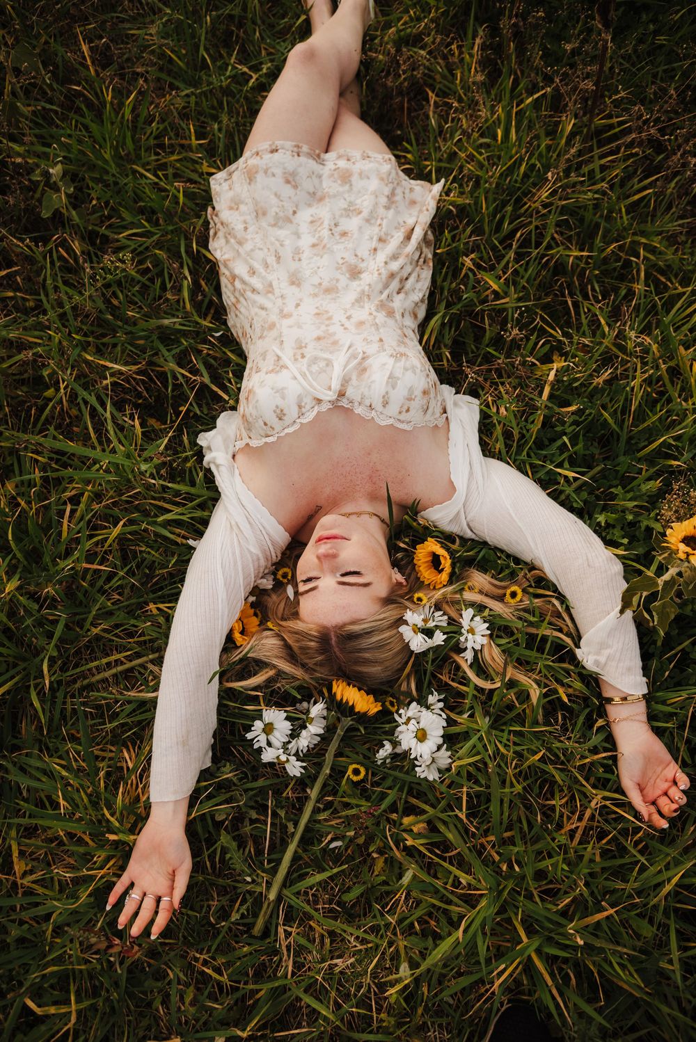 Person in white lace dress lying in grass surrounded by sunflowers and daisies in dreamy outdoor setting.