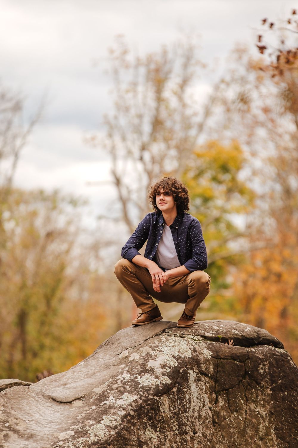 Person in casual attire sits perched on large boulder during autumn with fall foliage in background.
