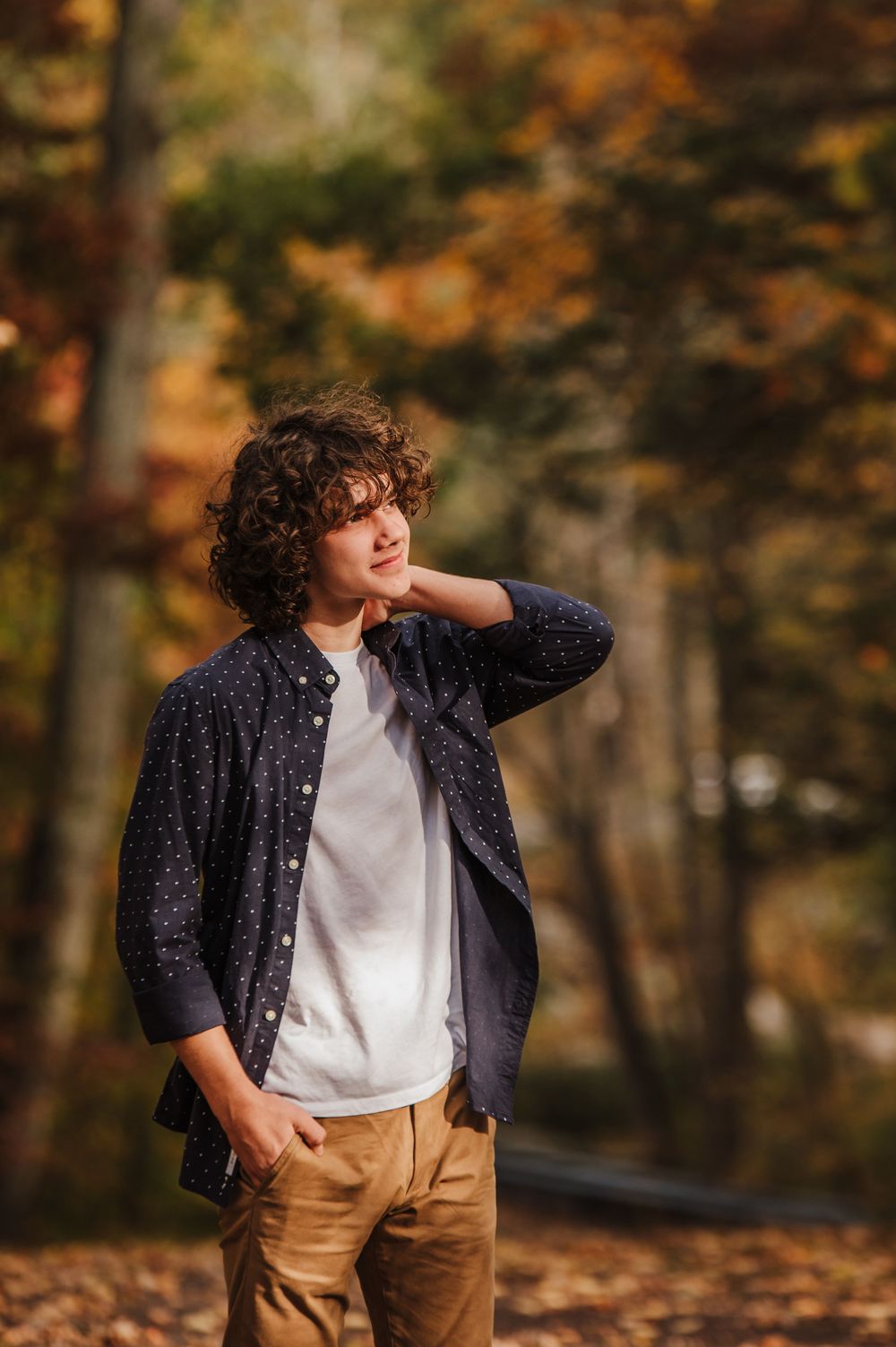 Person in navy polka dot shirt and khaki pants poses against autumn foliage backdrop in outdoor portrait session.