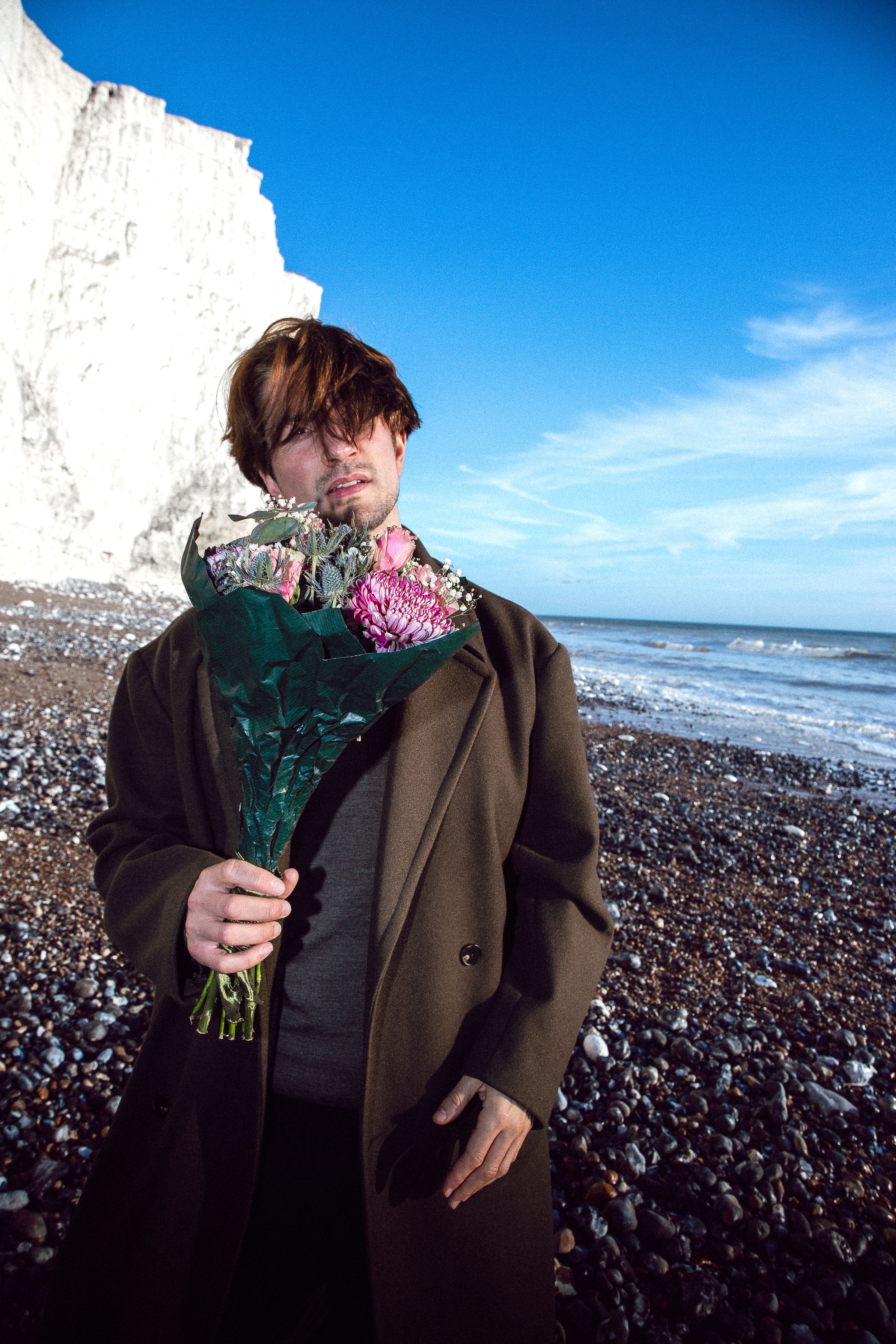 A figure in a brown coat stands on a rocky beach holding pink flowers against a bright blue sky and ocean backdrop.