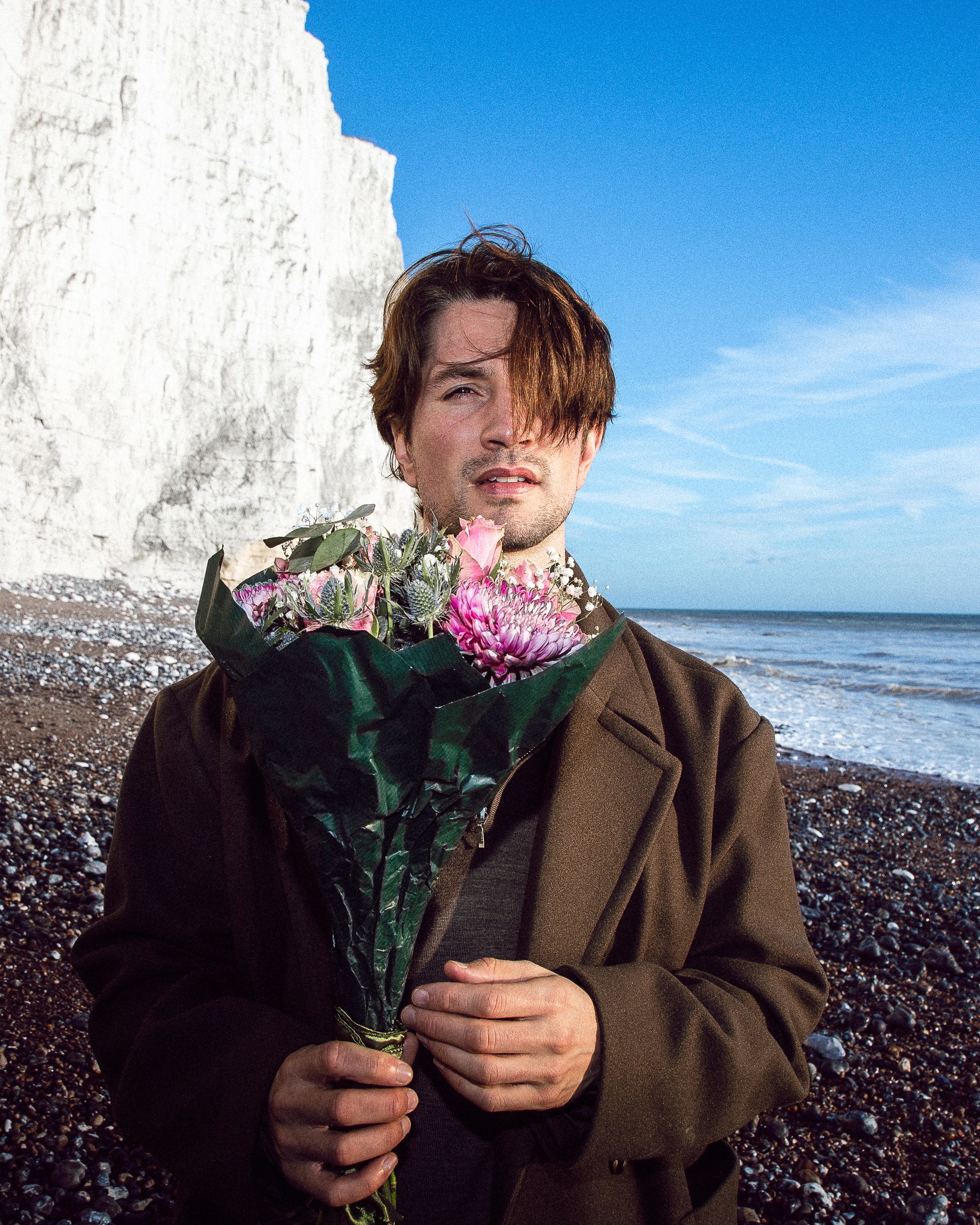 Person in brown coat holding pink flowers stands on rocky beach against bright blue sky and ocean.