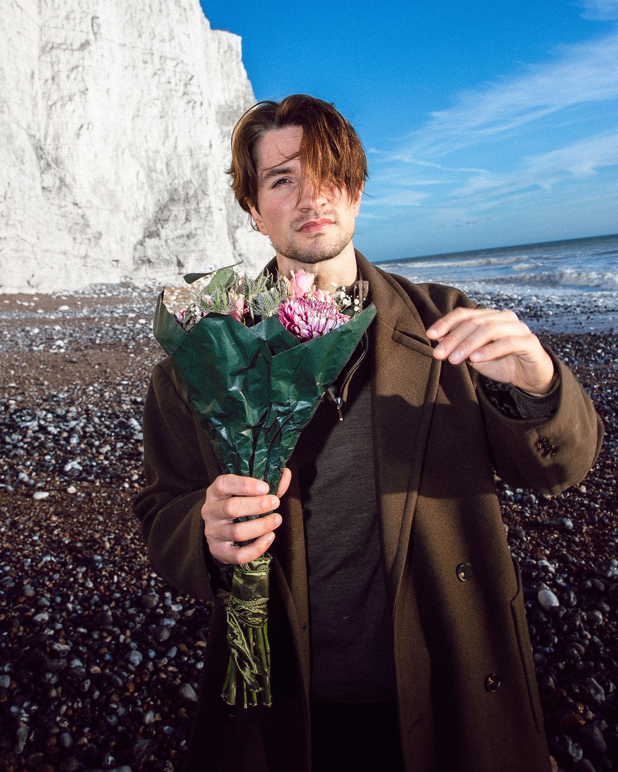 A person in a brown coat holds pink carnations against a backdrop of white cliffs and blue sky by the ocean.