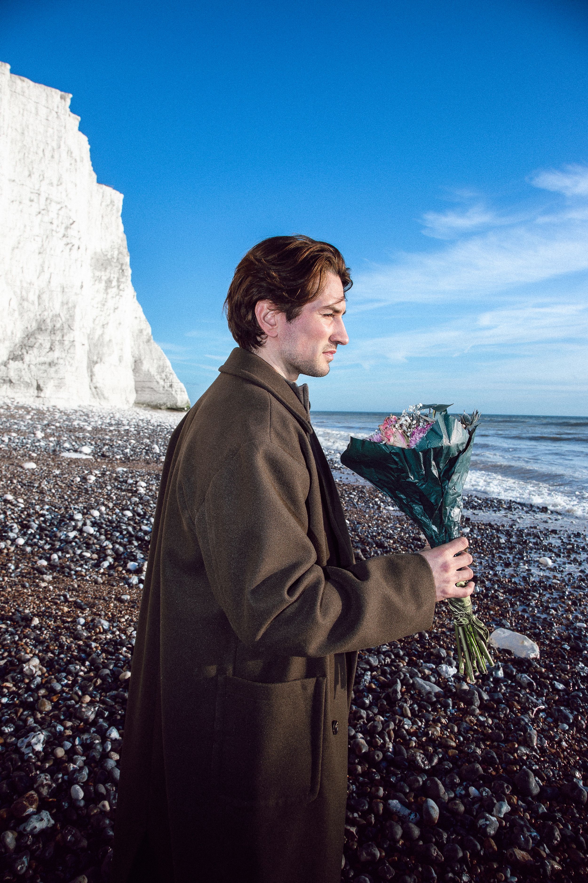 Two people stand on a rocky beach with white chalk cliffs in the background along the English coastline.