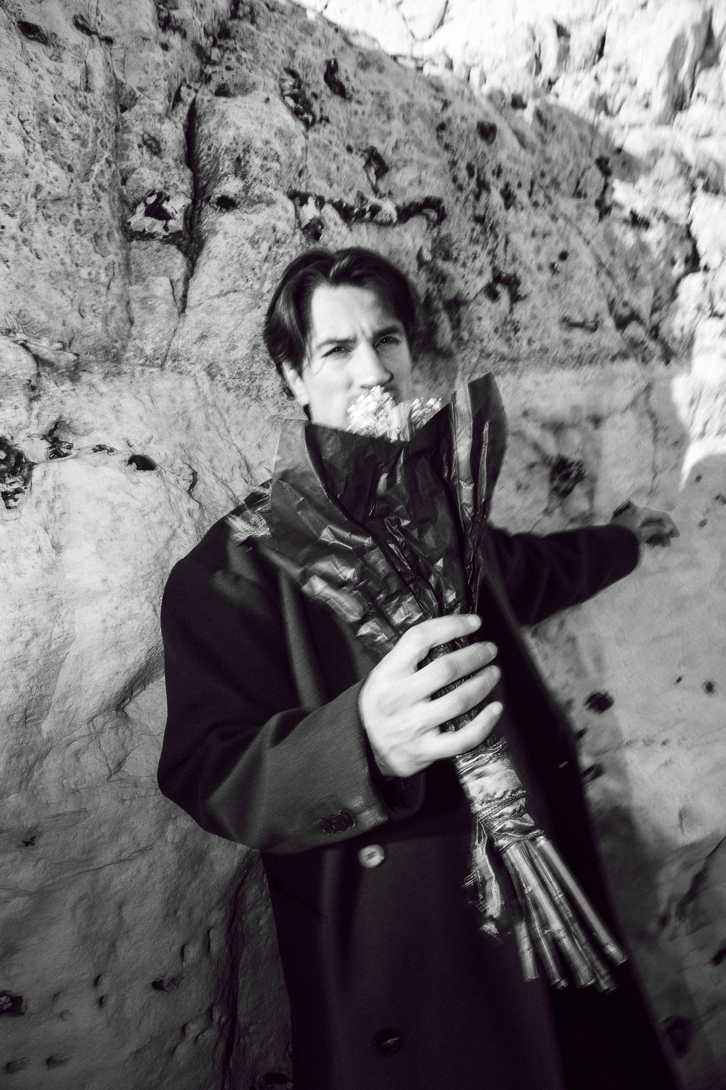 Black and white photo of someone in a dark coat holding a bundle of sticks against a stone wall background.