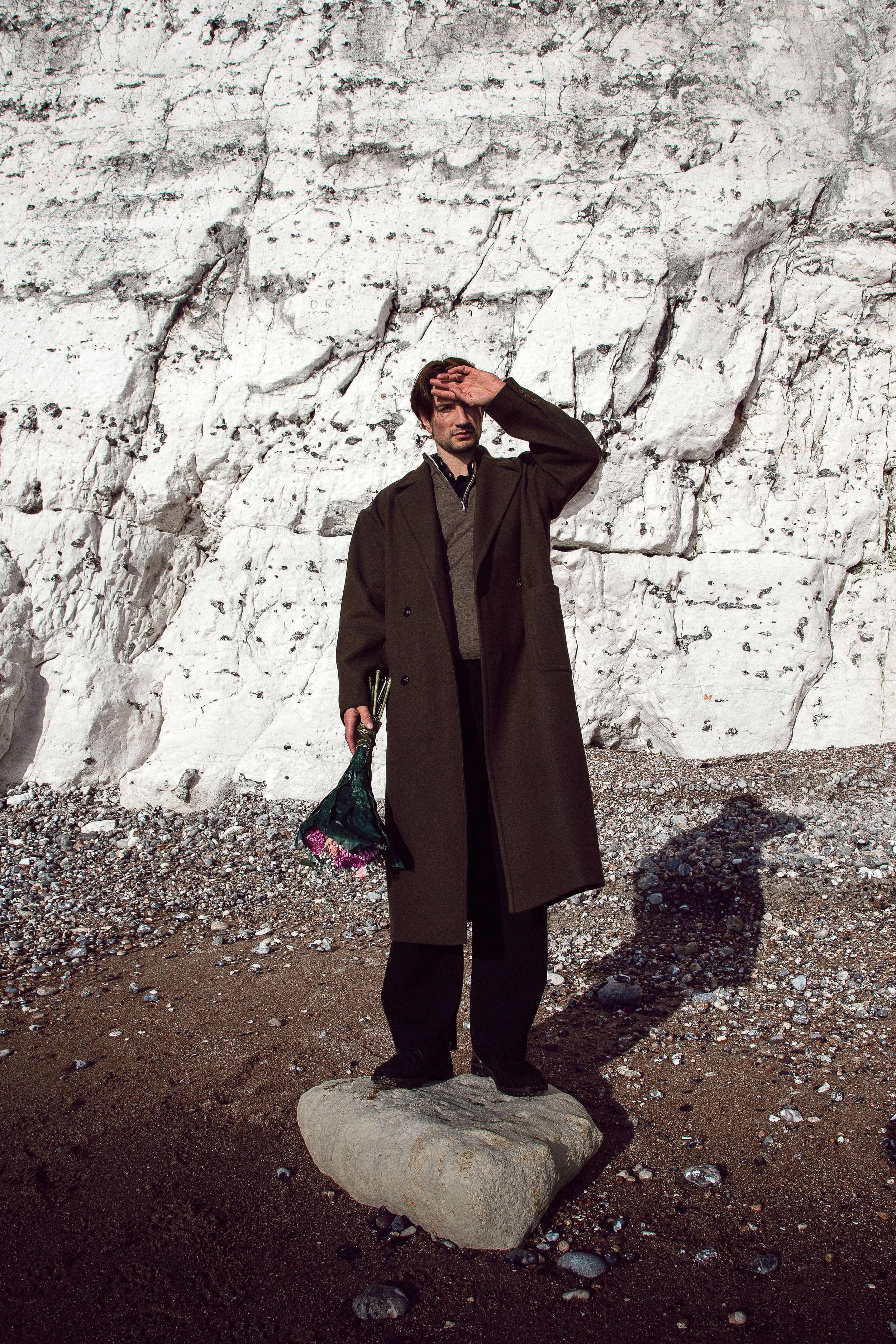 A person in a dark coat stands against a white stone wall in a sequence of dramatic poses.