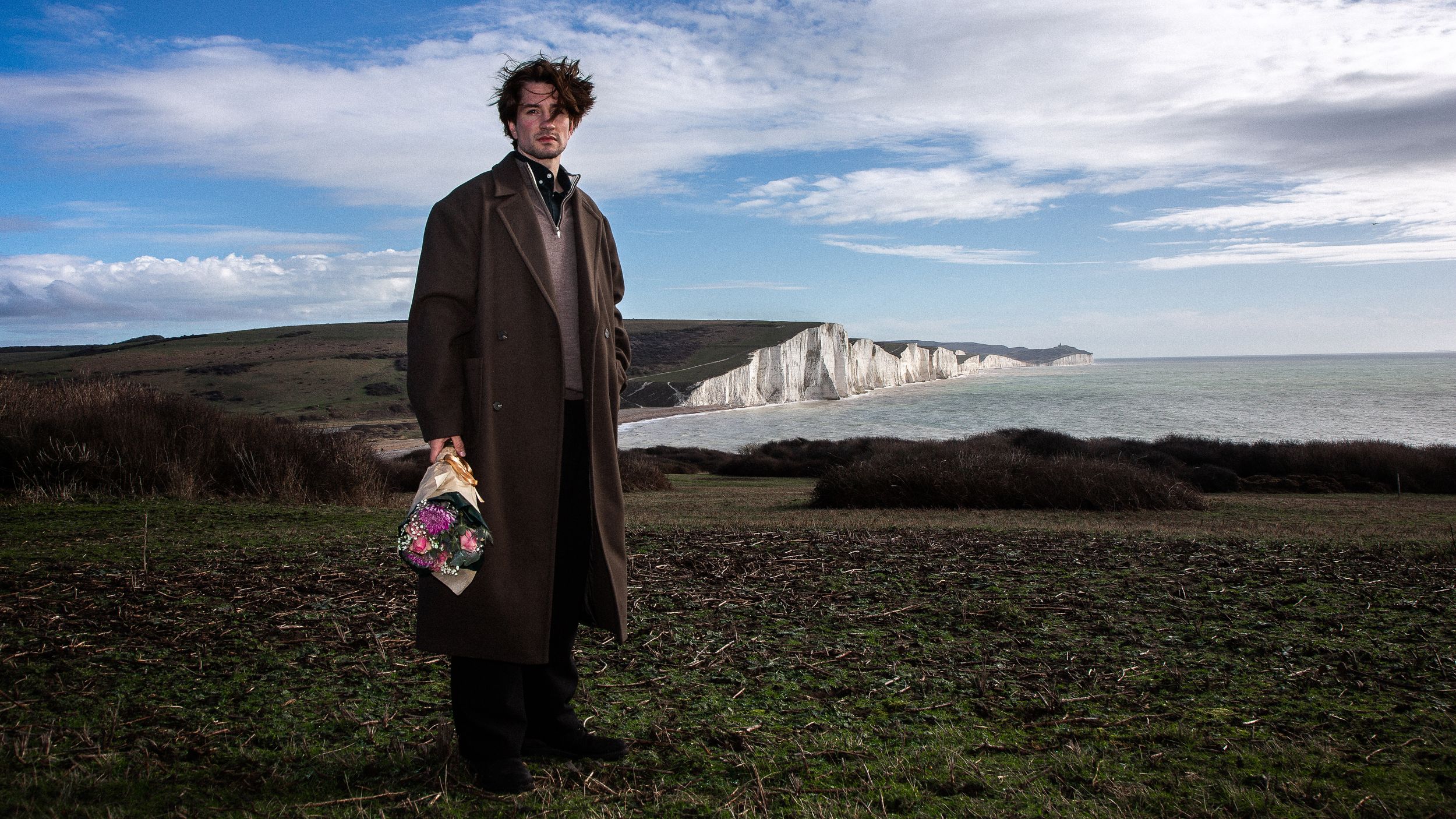 A person in a brown coat stands on a grassy hilltop overlooking white coastal cliffs and ocean on a sunny day.