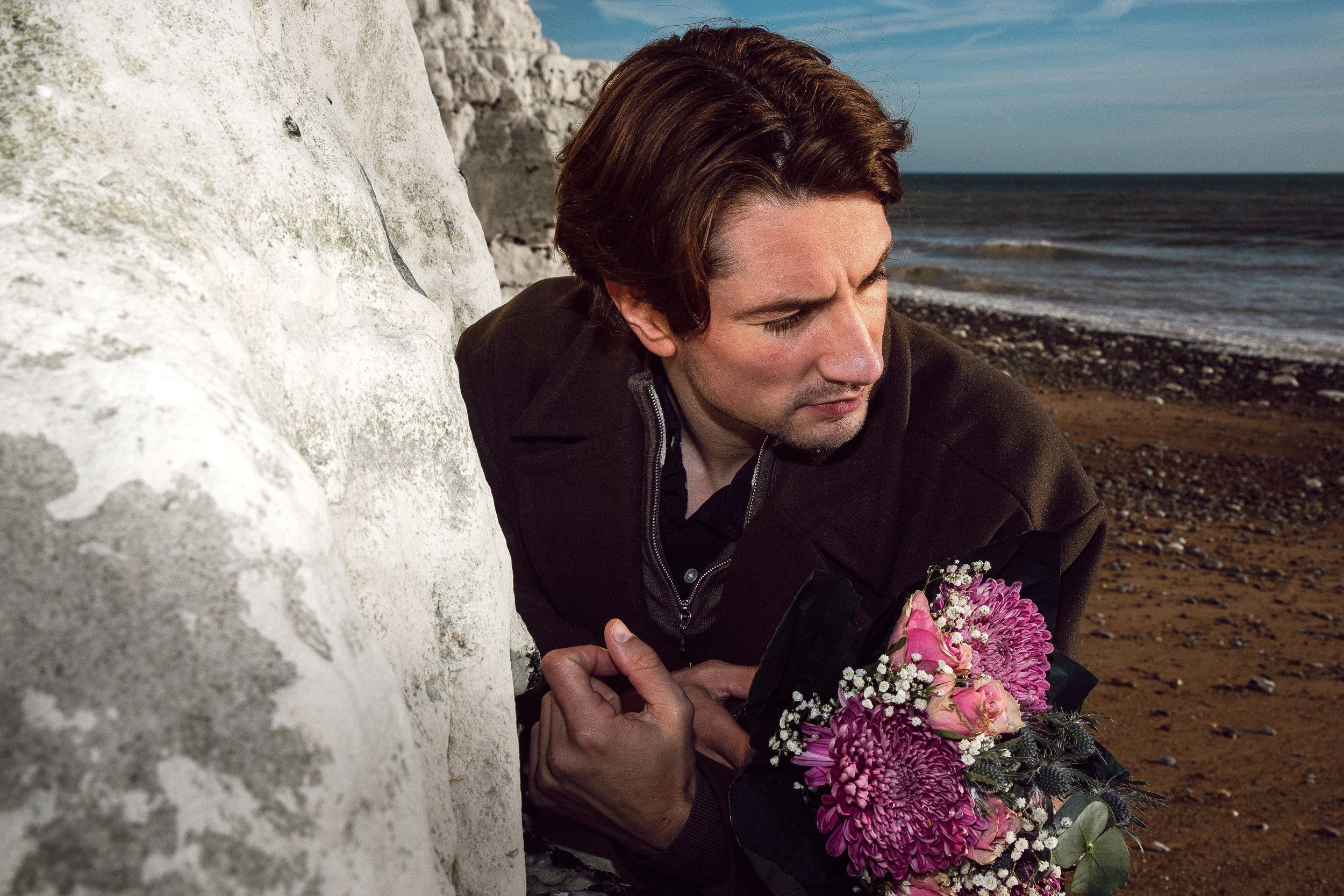 Person holding pink flowers poses near white cliffs overlooking a coastal beach view.