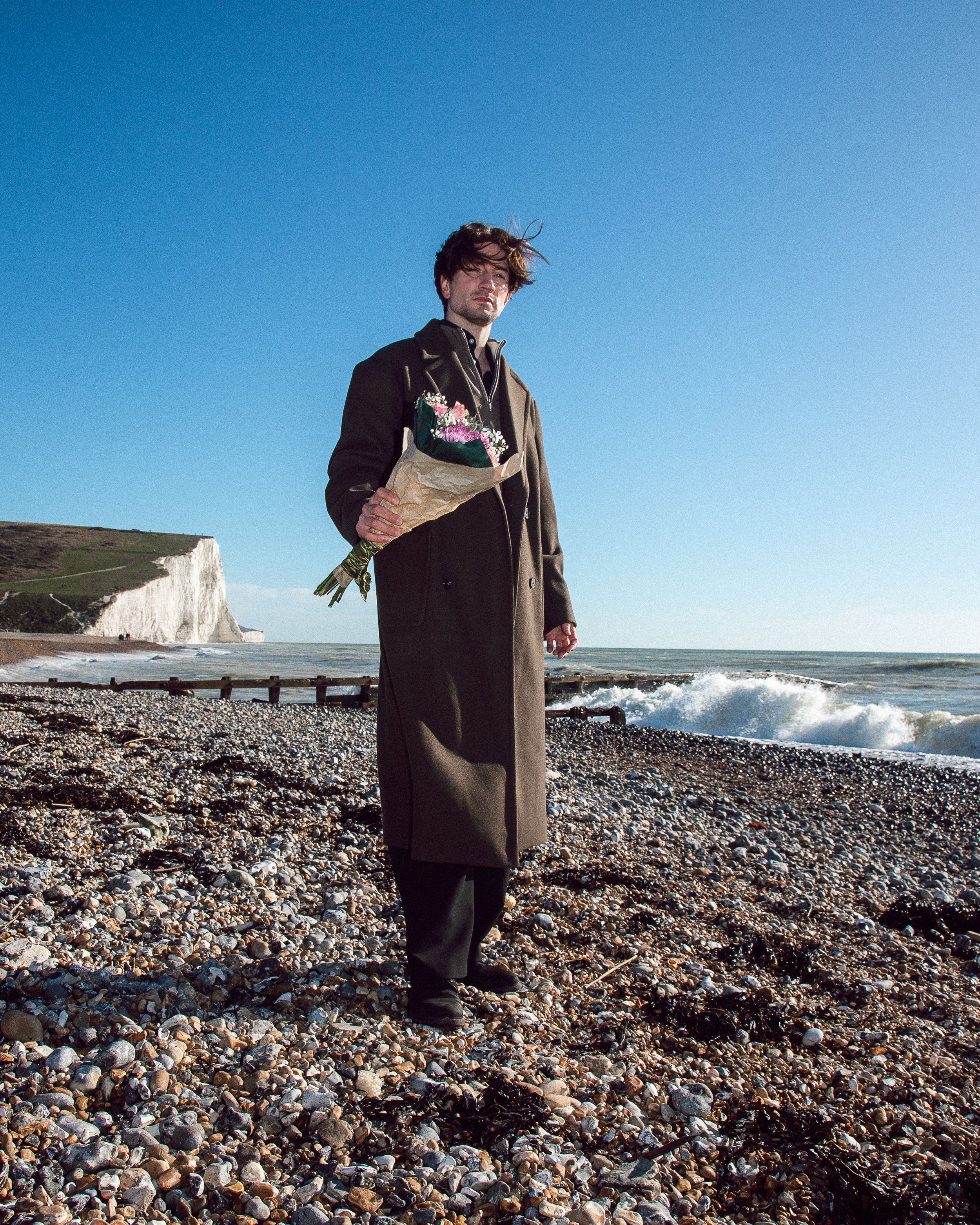 A figure stands on a rocky beach with white cliffs and crashing waves at Seven Sisters, wearing a long dark coat.