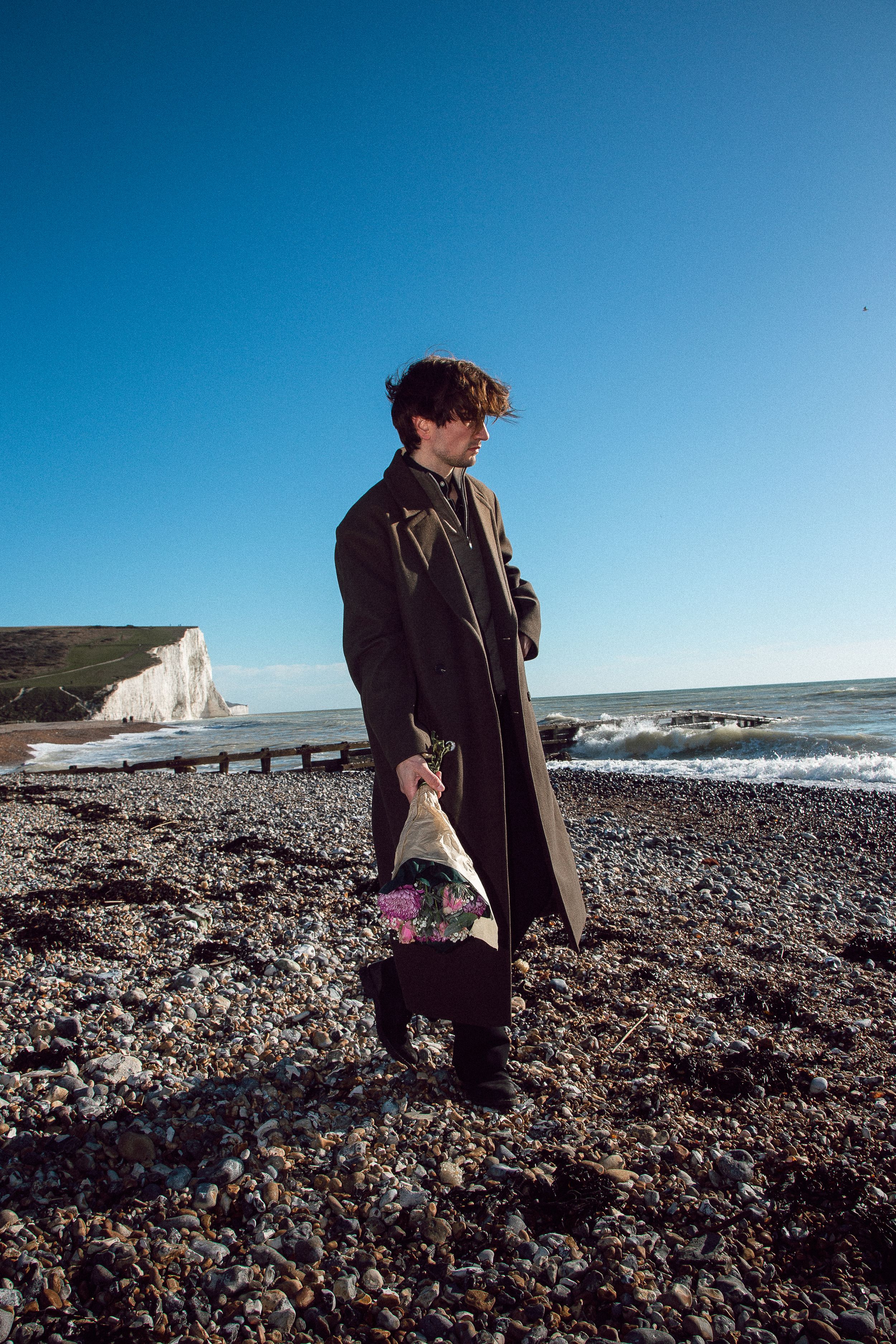 Person walking along rocky beach with white cliffs and ocean waves in background on sunny day.