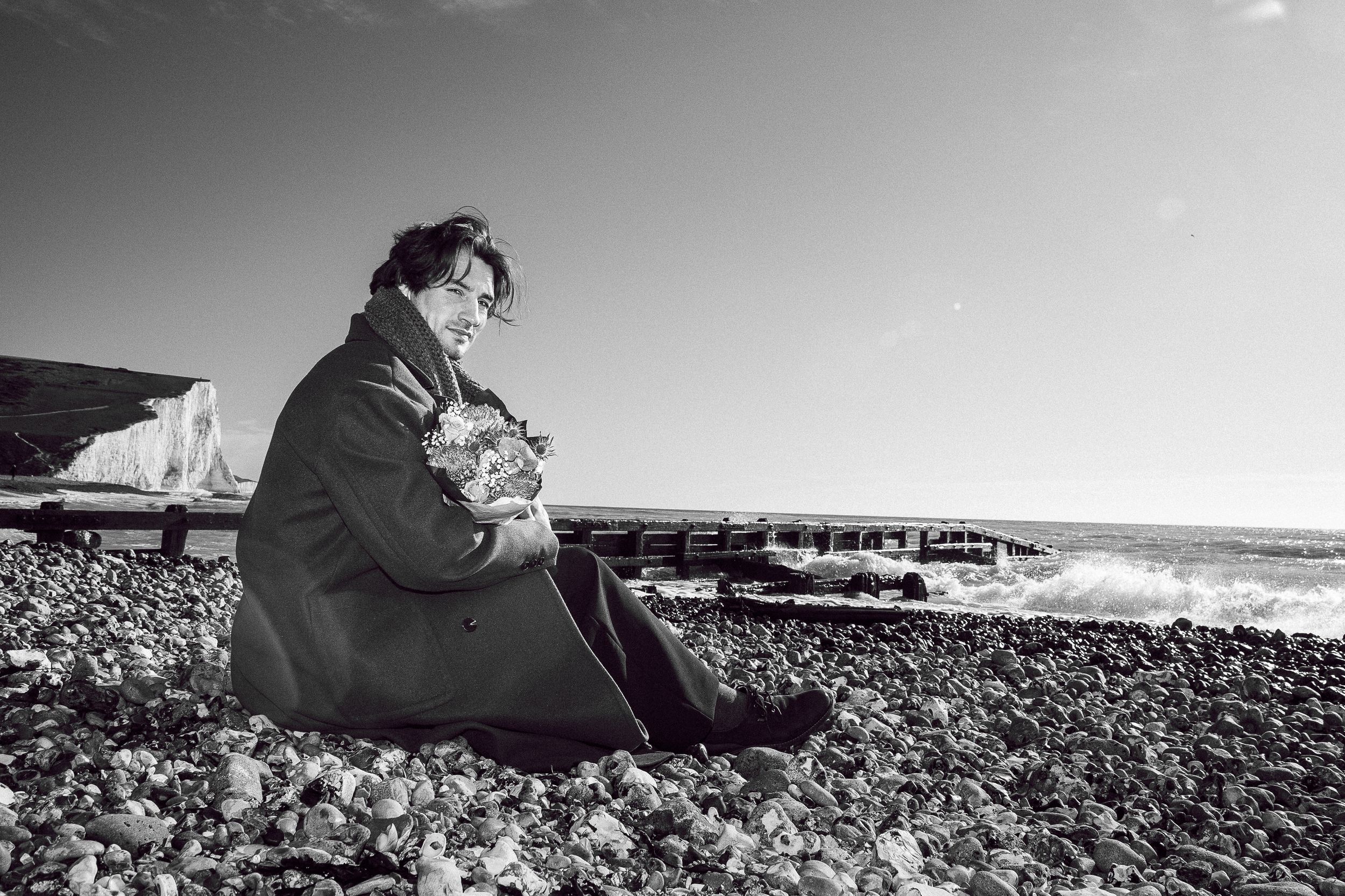 A series of black and white beach photographs showing intimate moments between two people on a pebbled shoreline.