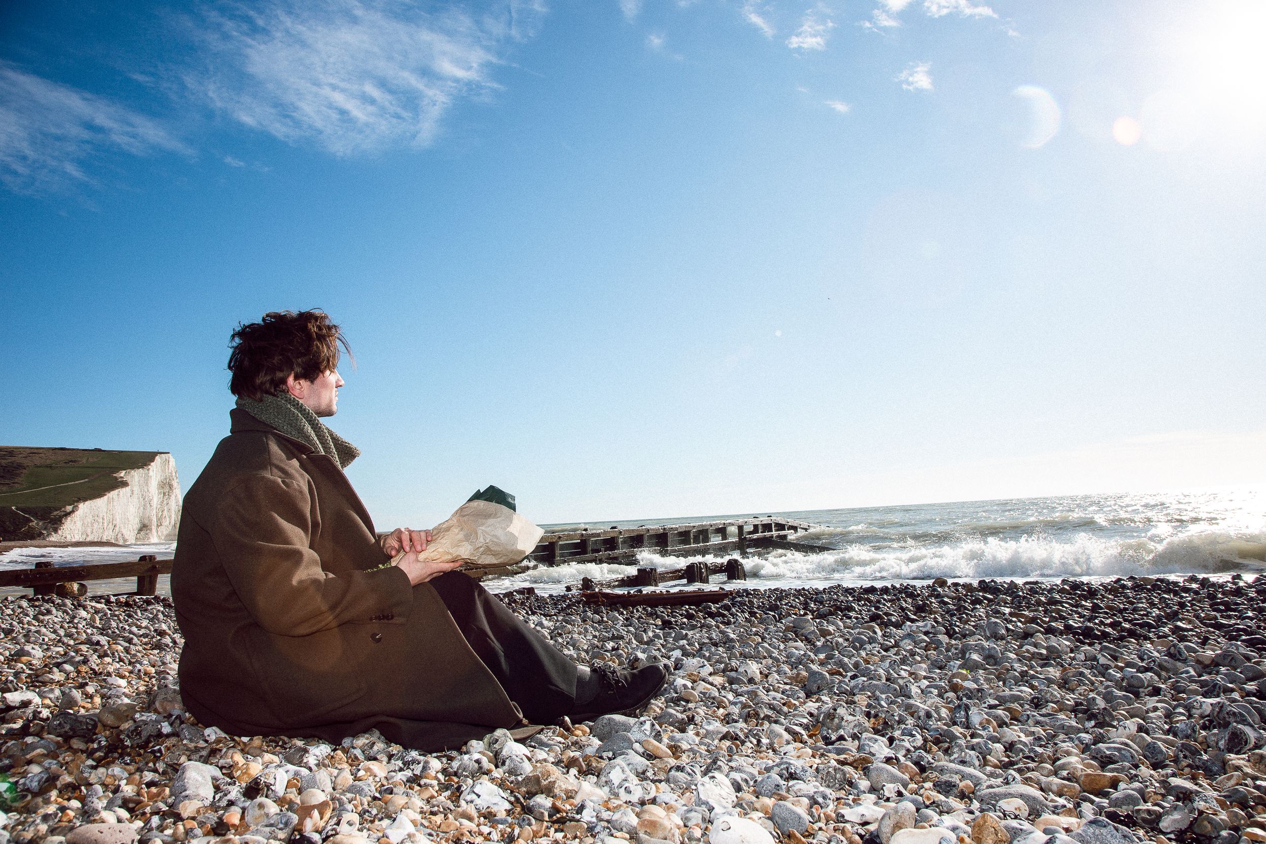 Person sitting on a rocky beach looking out at the ocean waves and blue sky on a sunny day.