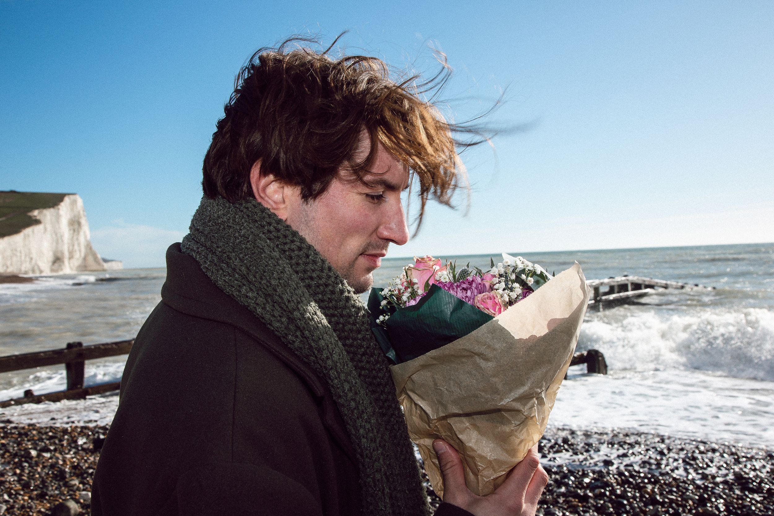 Someone in a dark coat holds a paper bag while standing on a windy beach with white cliffs visible in the background.