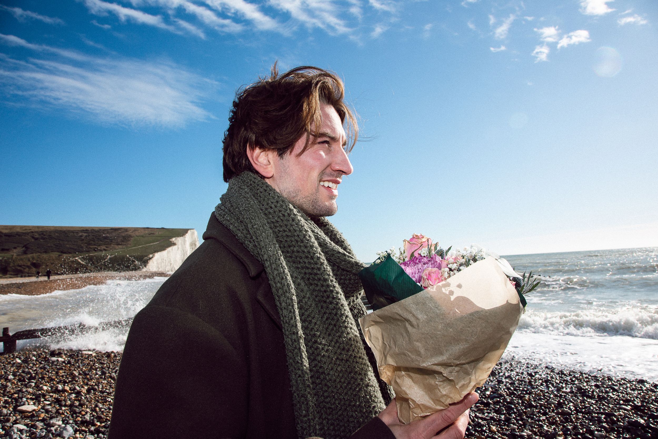 A person wearing a dark coat and scarf sits on a rocky beach holding flowers while looking at the ocean.