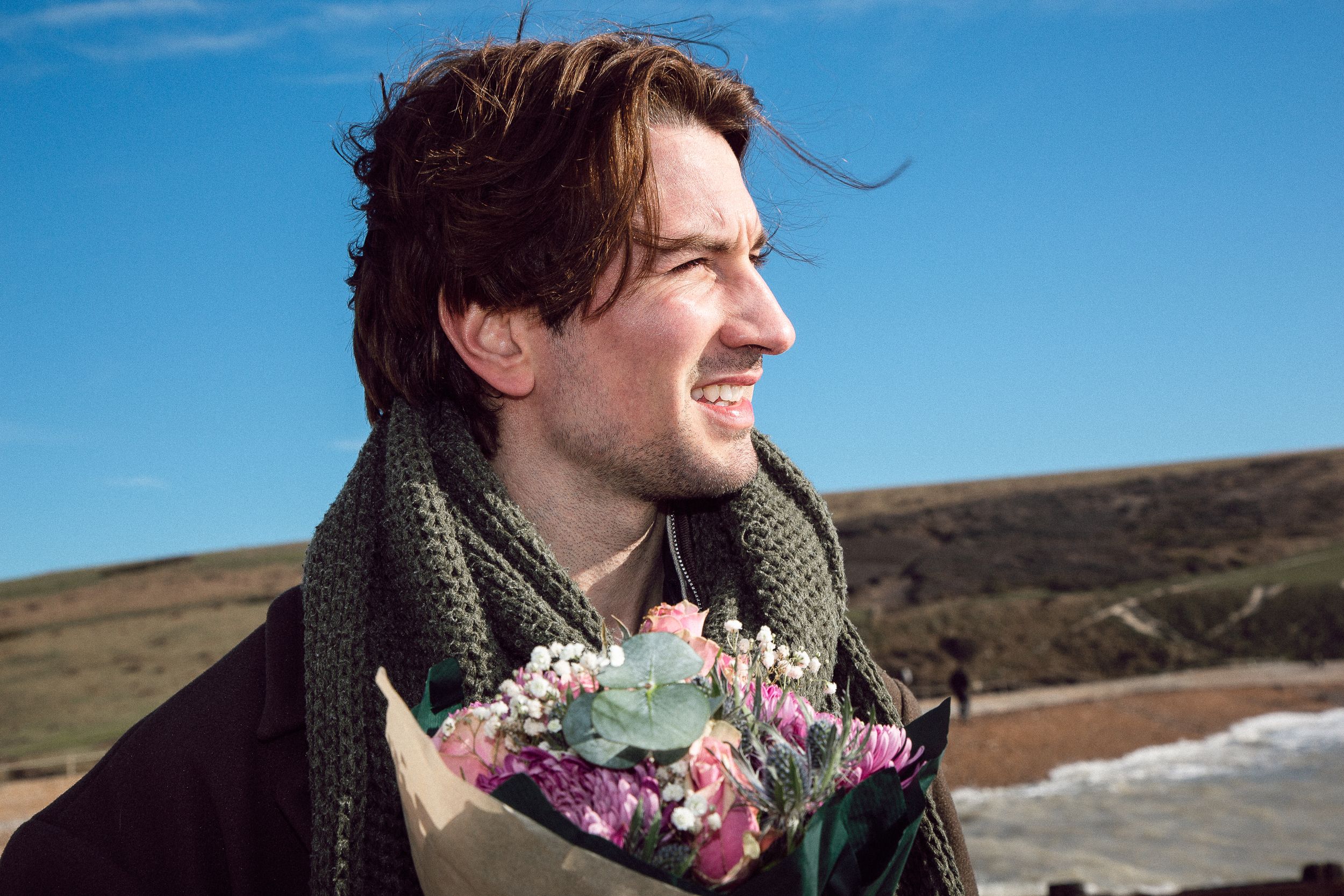 A person in a dark scarf smiles while holding a bouquet of flowers against a bright blue sky and coastal landscape.