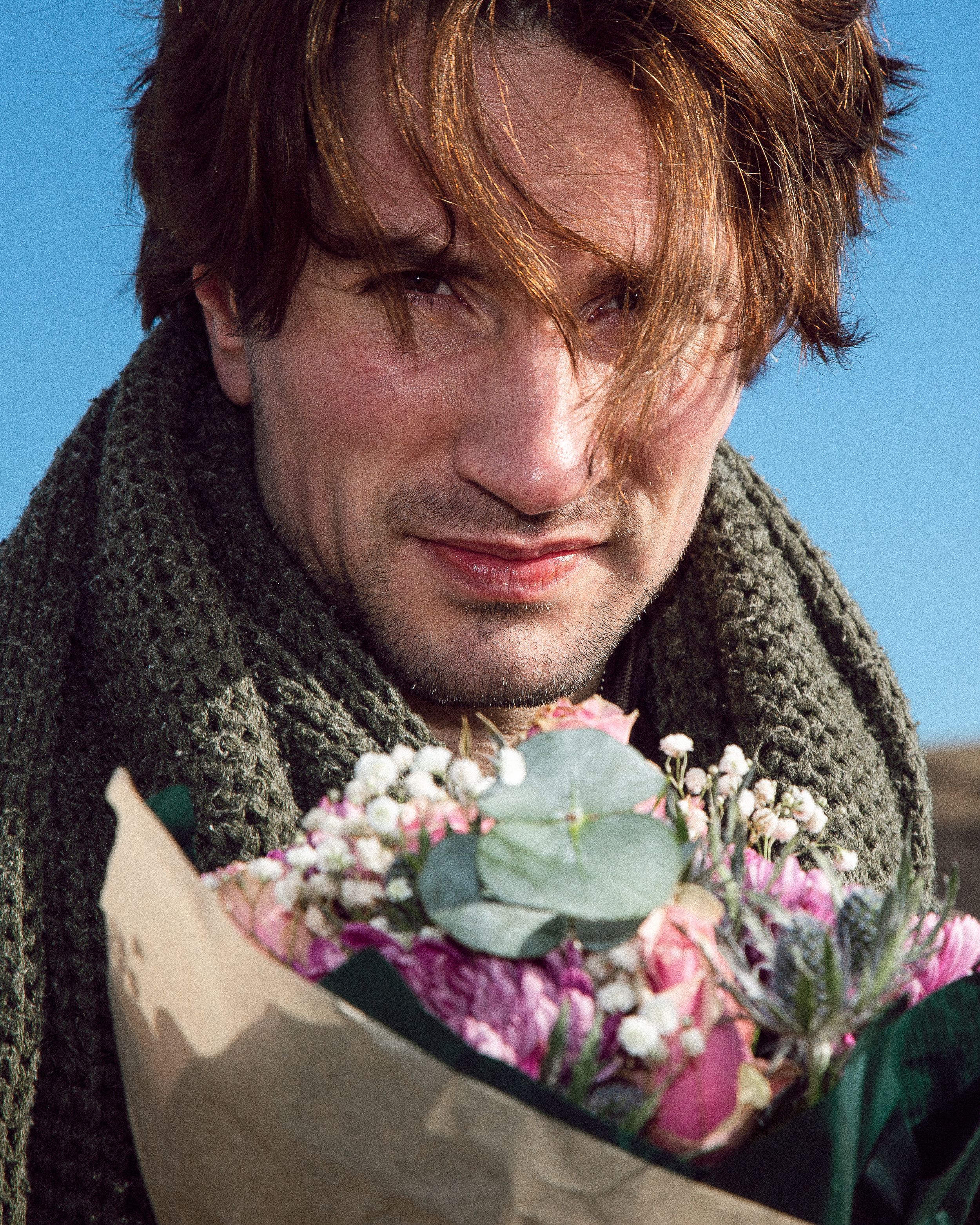 A person in a dark coat smells a bouquet of pink and white flowers against a bright blue sky background.