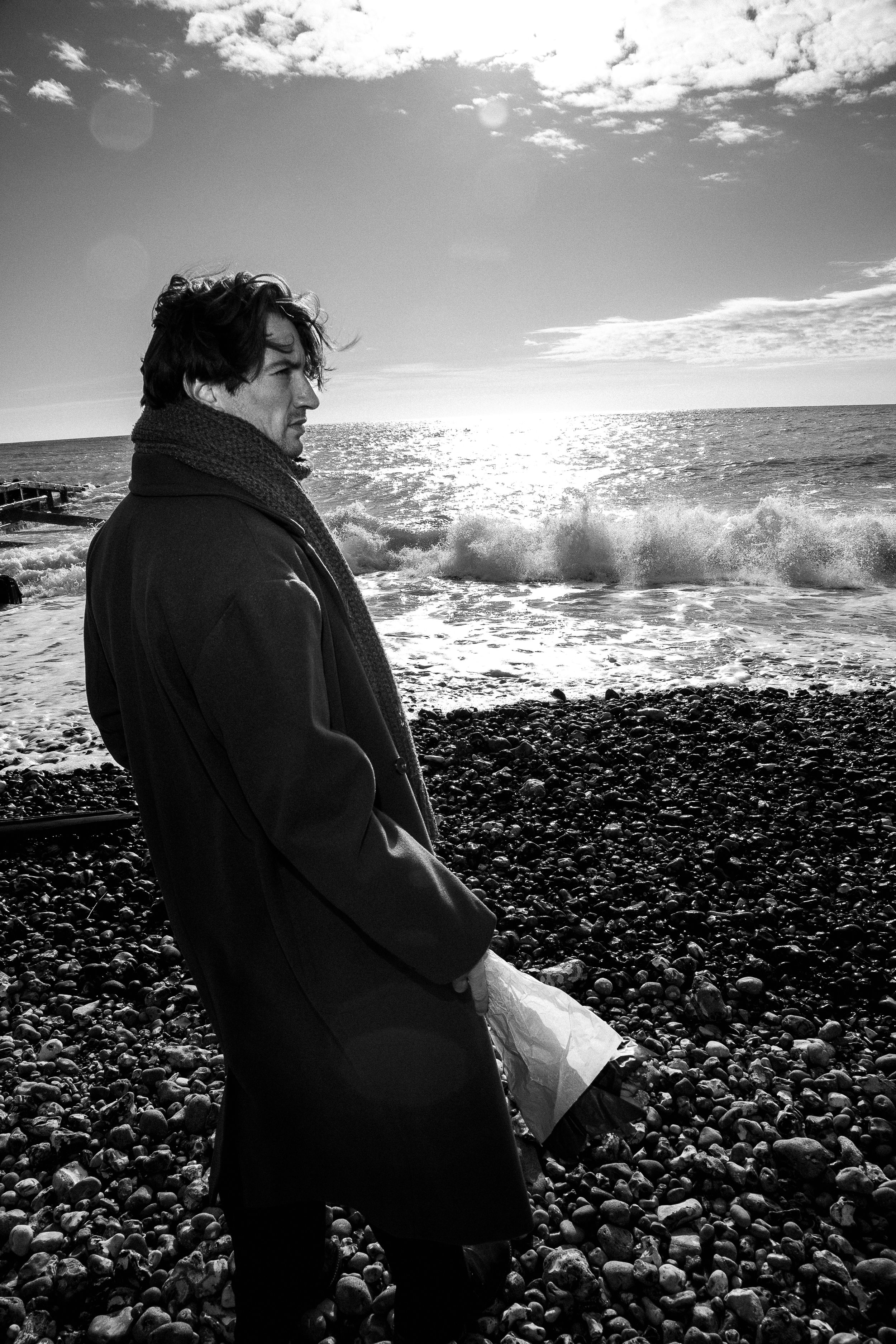 A person in black stands on a rocky beach as waves crash against the shore in dramatic black and white photography.