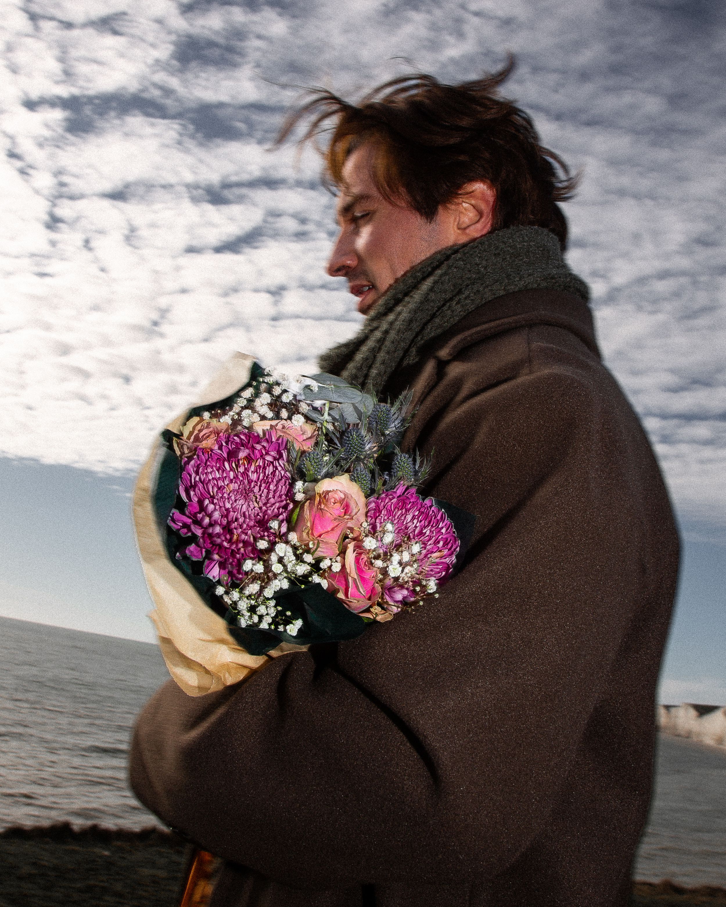 A person in a brown coat holds a vibrant bouquet of pink flowers against a dramatic cloudy sky by the ocean.