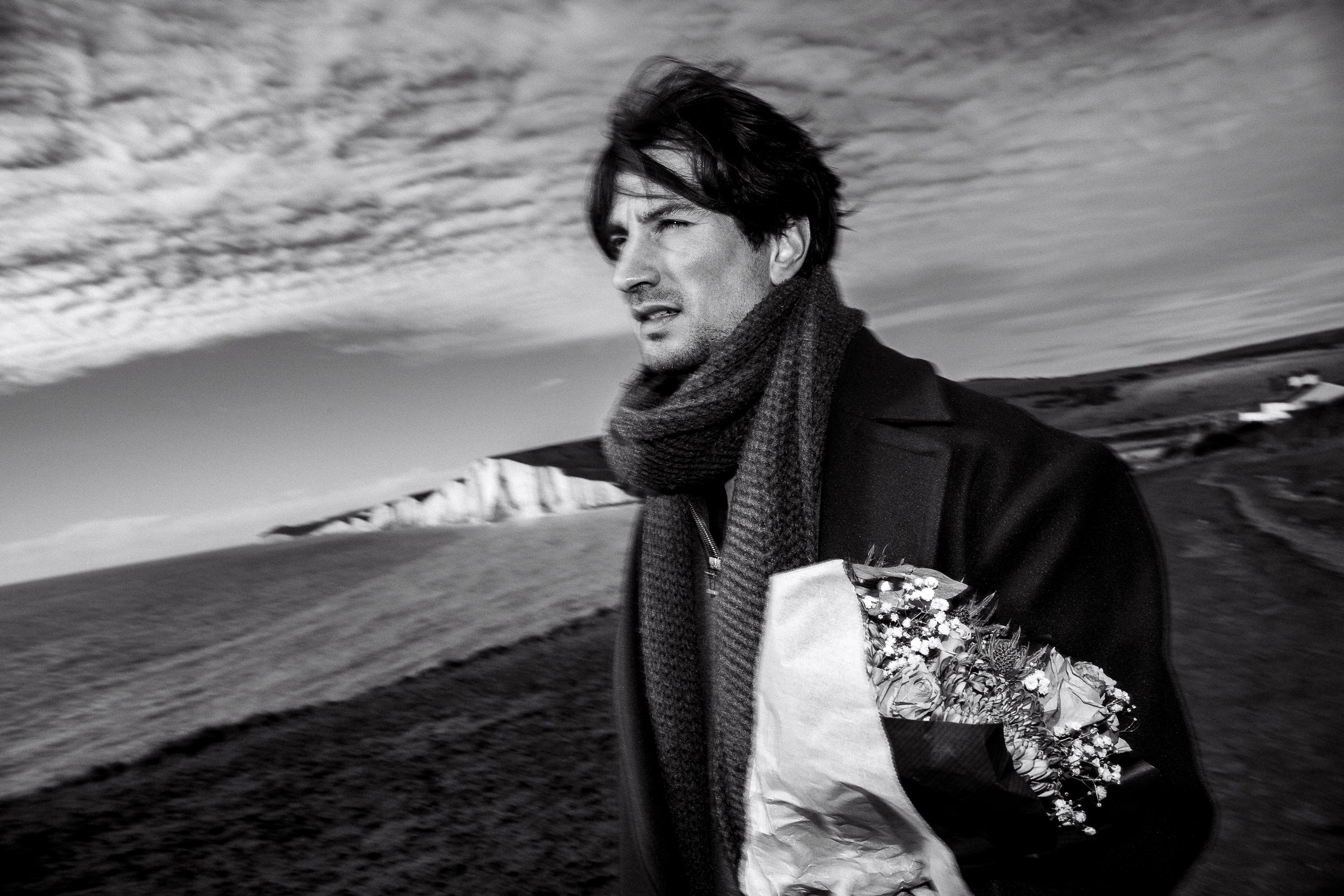 Black and white photo of a person wearing a scarf standing by the ocean coast under dramatic cloudy skies.