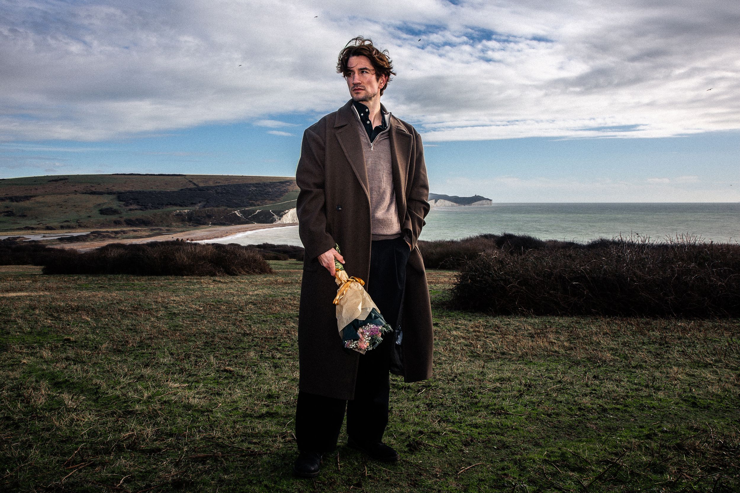 Figure in a long brown coat stands on coastal cliffs holding flowers against a cloudy sky.
