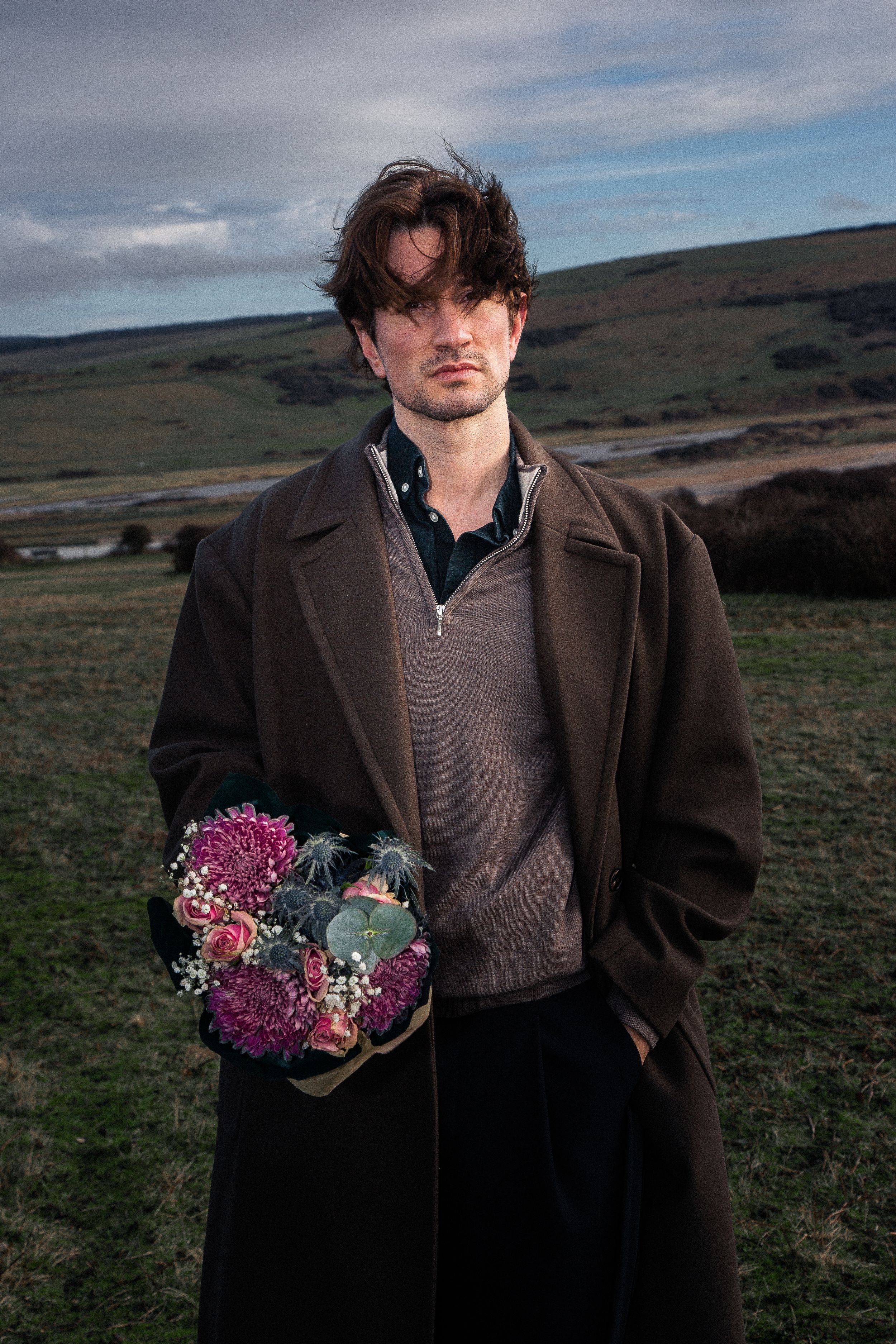 Person in dark coat holds pink and blue flower arrangement while standing on windswept coastal hillside.