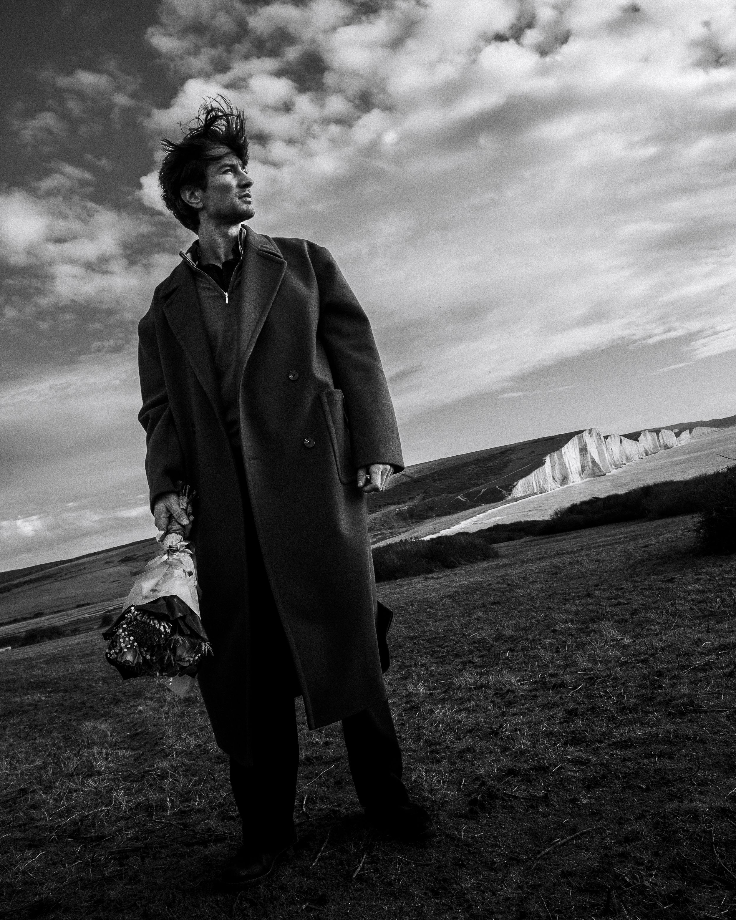 A person in a dark coat stands against a dramatic cloudy sky on a grassy hillside in black and white.